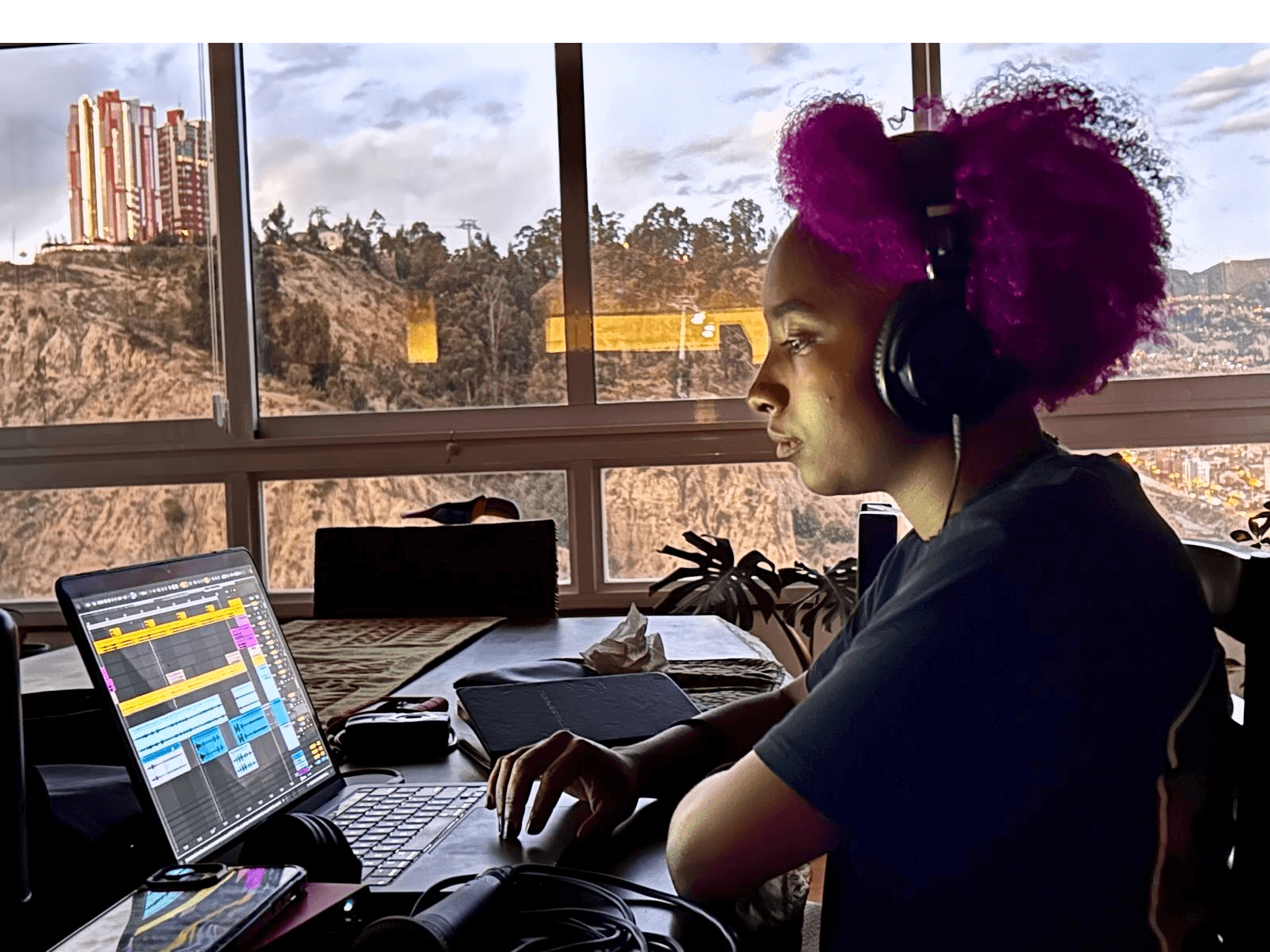Young woman with pink curly hair wearing headphones, working on a laptop in a room with large window and outdoor view.