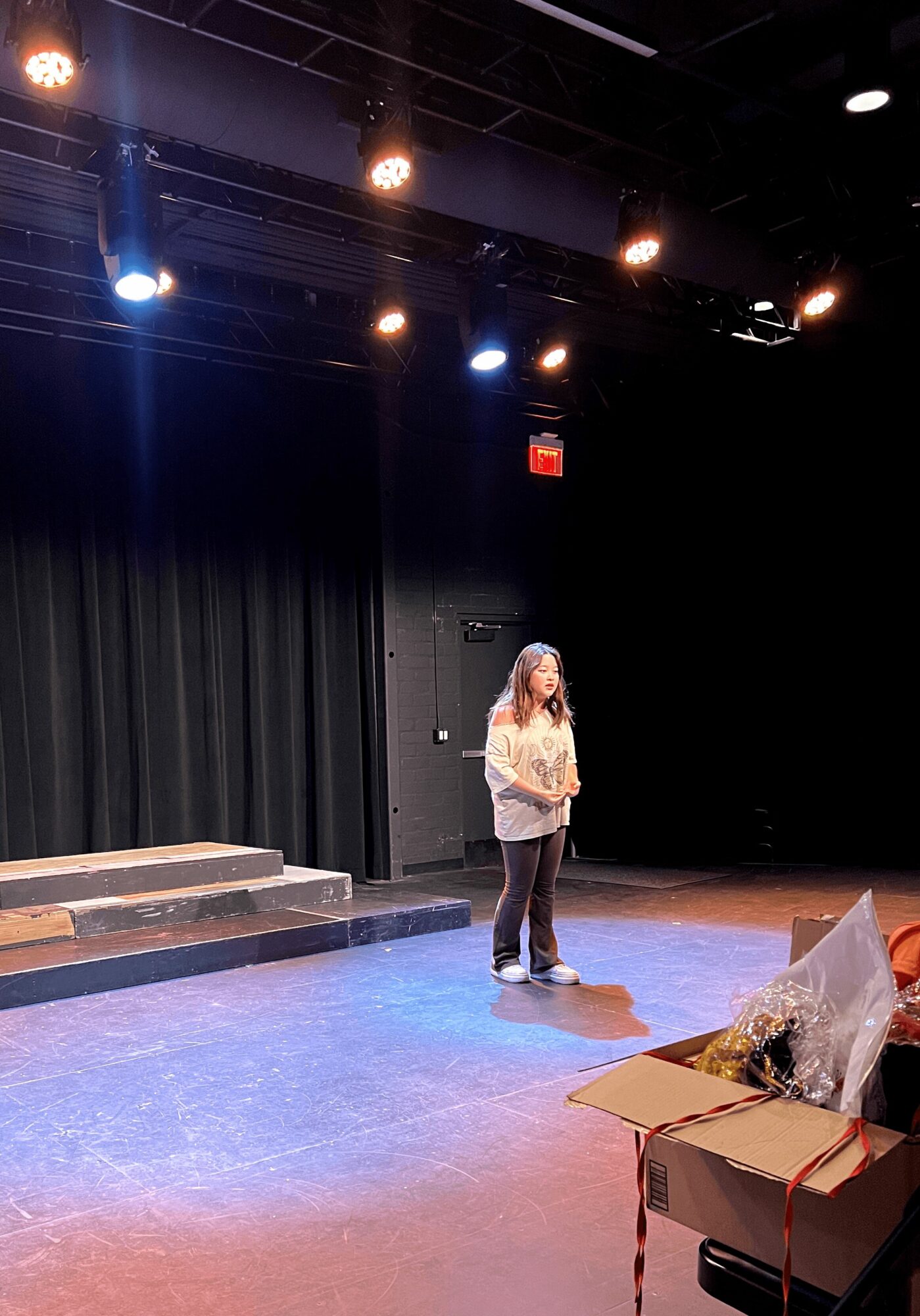 Person standing on stage with black curtain backdrop, stage lights overhead, and a table with items in foreground.