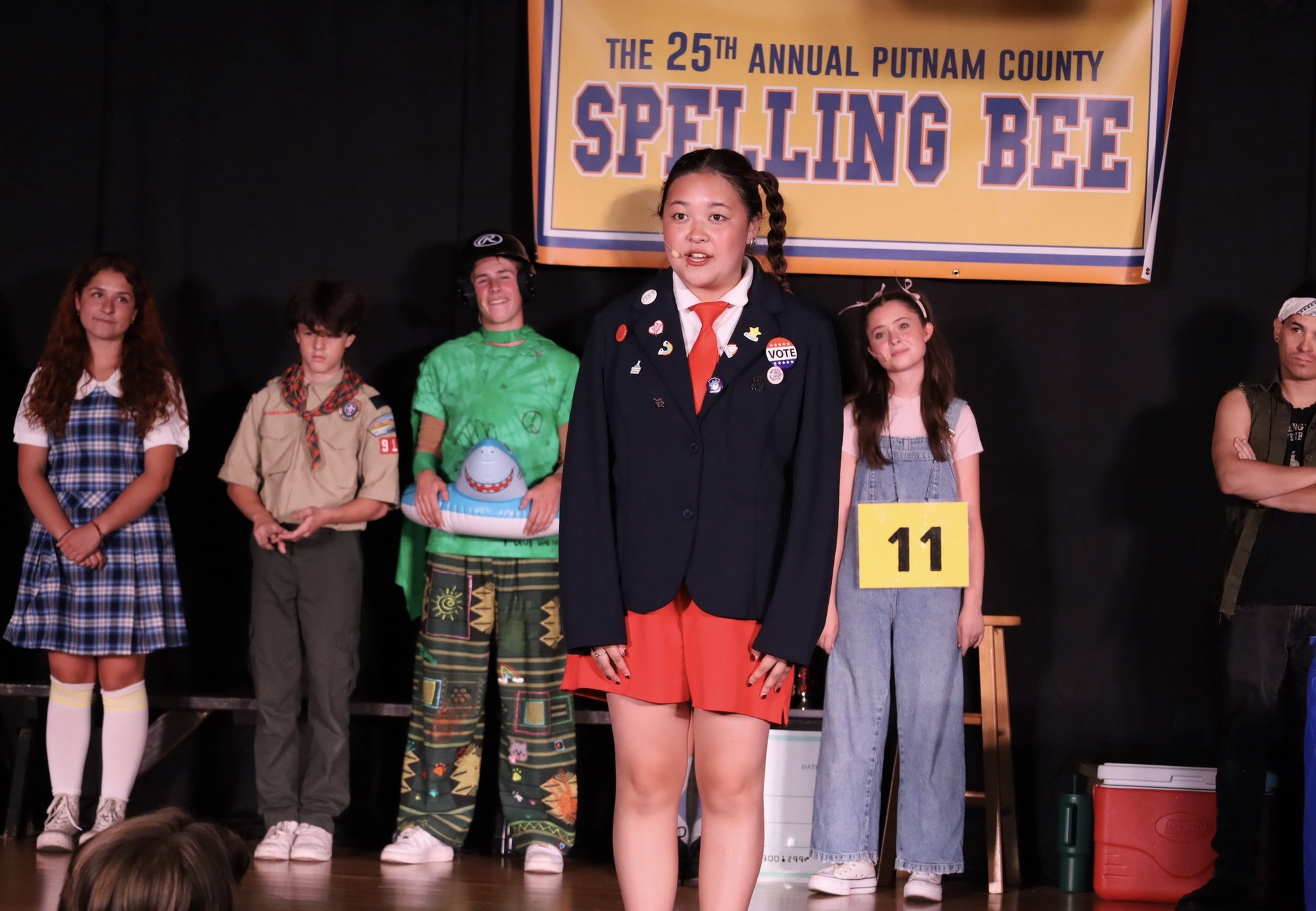 Girl in uniform speaking on stage at spelling bee event with children in background and banner above.