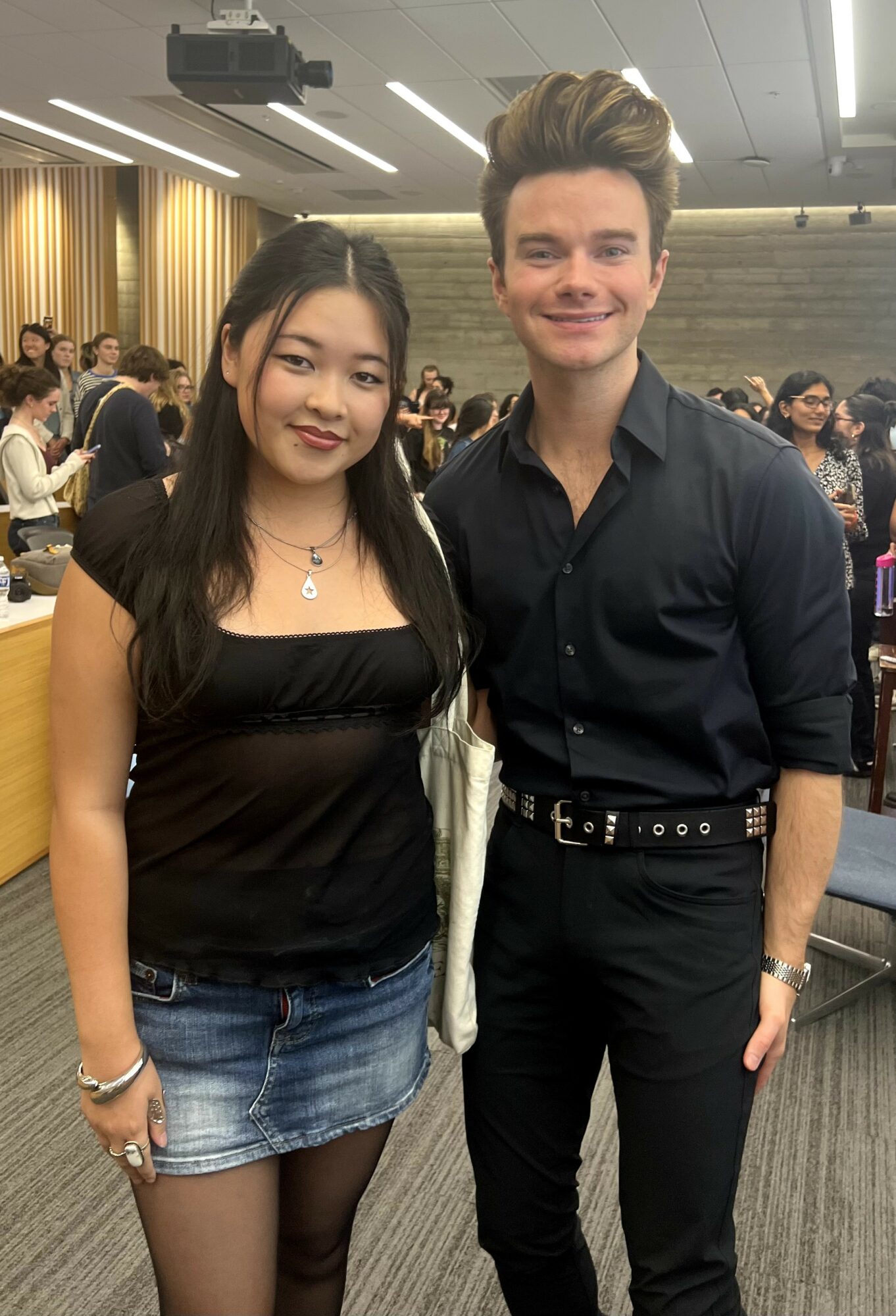 A young woman and a young man stand together in a conference room, smiling at the camera.