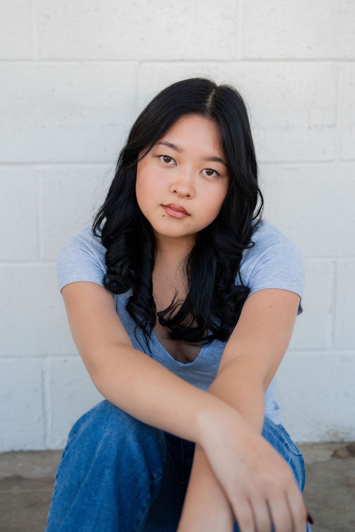 Young woman with long black hair wearing a light blue shirt, sitting against a white brick wall.