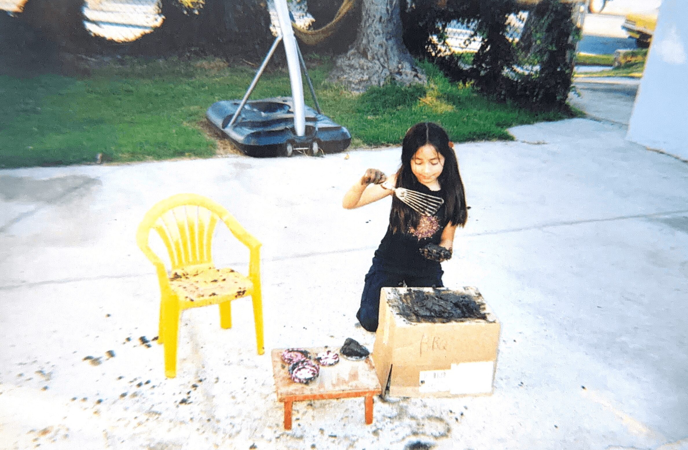 Girl with long dark hair standing outdoors next to a large box with a cake, yellow chair nearby, on concrete surface.