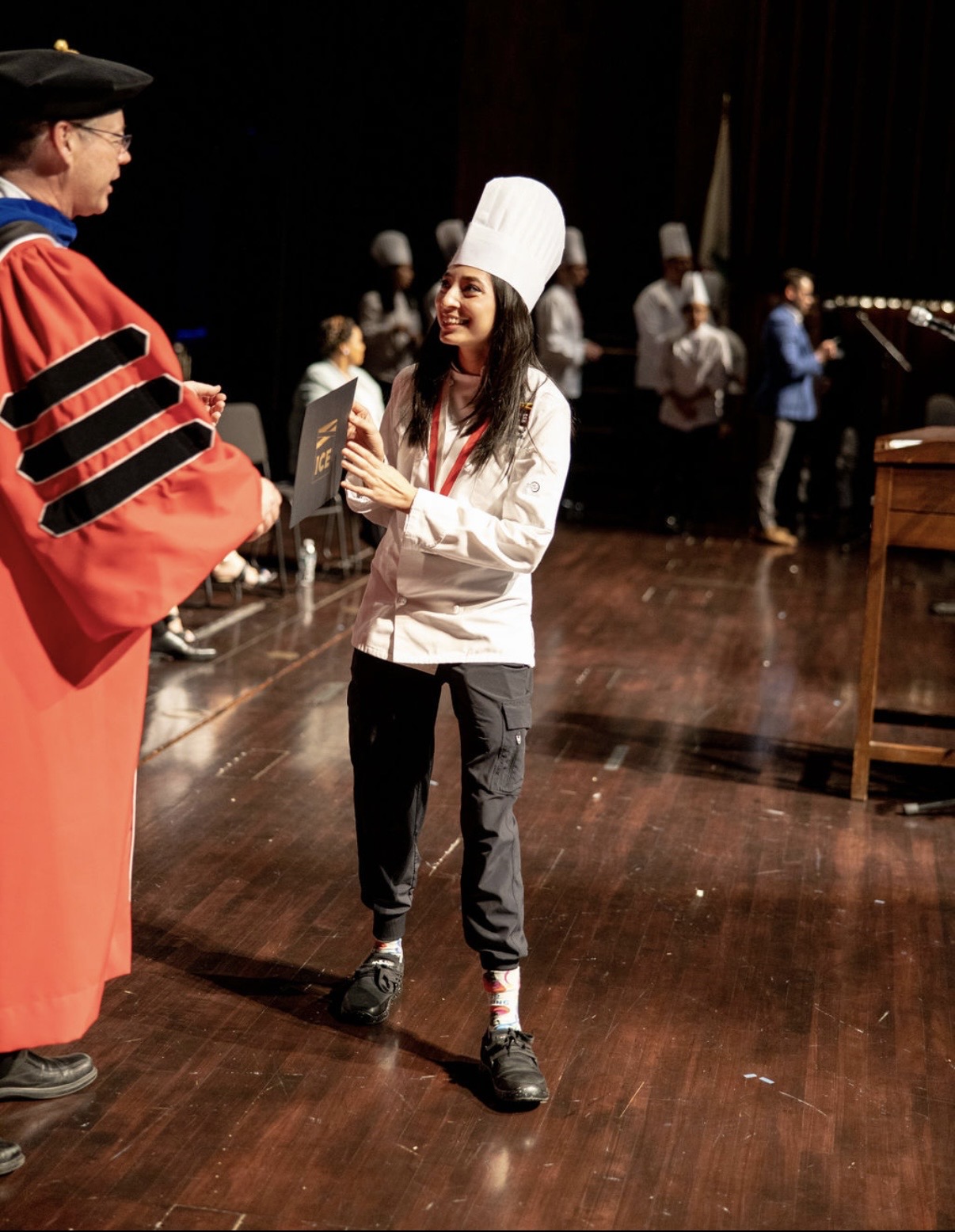 Young woman in chef's hat and white jacket receiving diploma from person in academic gown on stage.