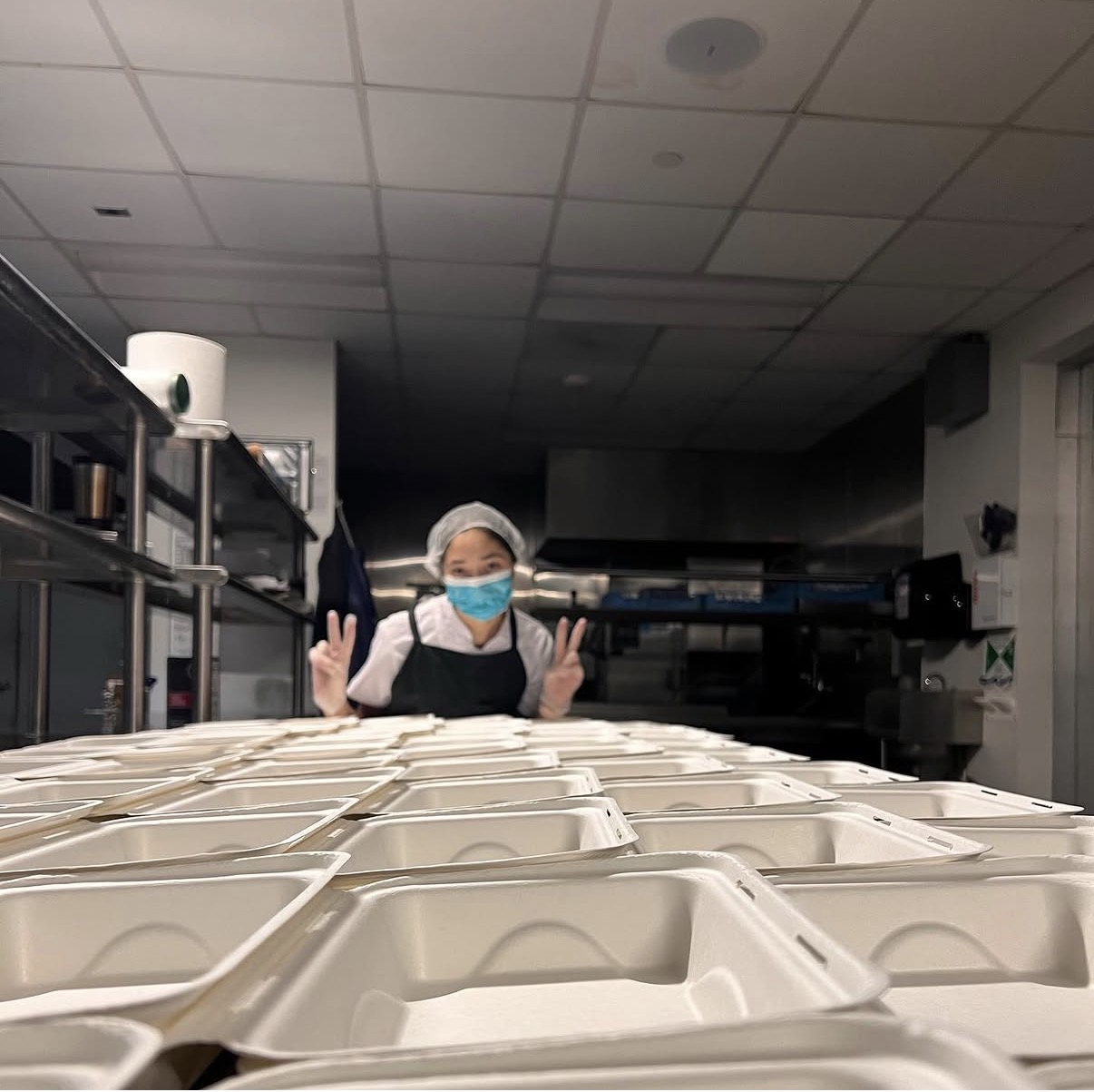 Person wearing a mask and hairnet making peace signs behind a row of empty food containers.