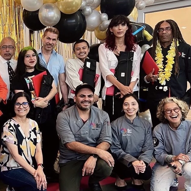 Group of ten people celebrating graduation, some holding diplomas, in front of black, white, and gold balloons.