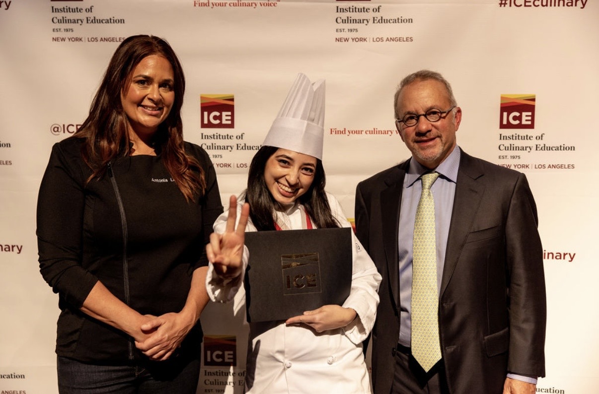 Three people standing in front of a backdrop, one in a chef's hat and uniform, two in formal attire, smiling.