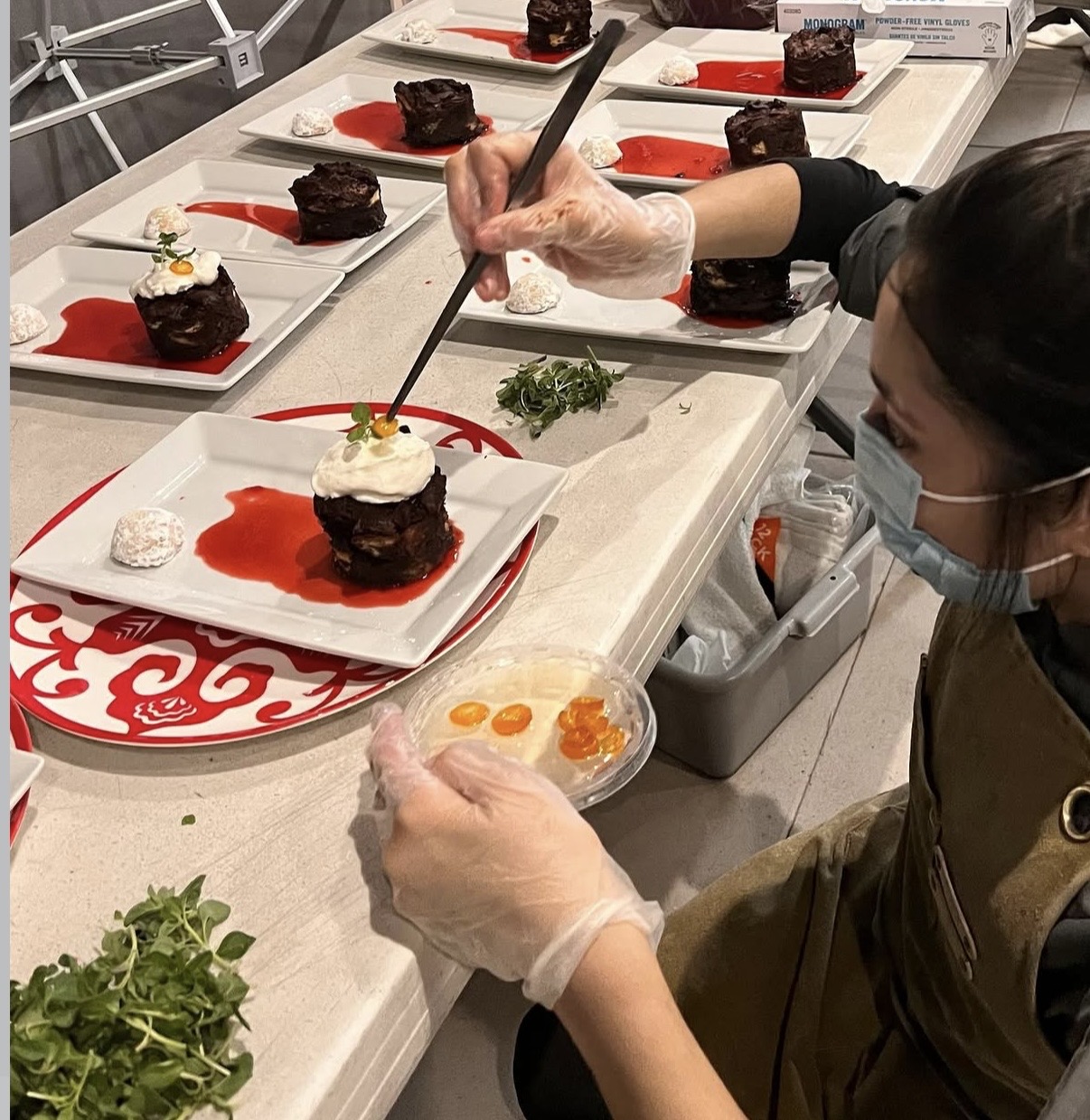 Person decorating plated desserts with a brush, wearing gloves and a face mask, on a white counter with multiple desserts.