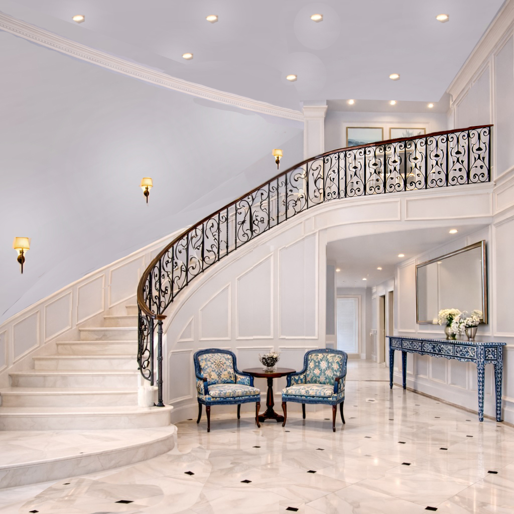 Elegant staircase with ornate railing, two chairs, a small table, and a console table with flowers in a spacious, well-lit interior.