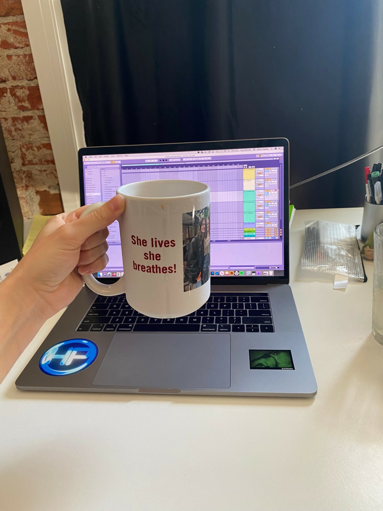 Hand holding a mug with text in front of a laptop on a desk, music production software open on screen.