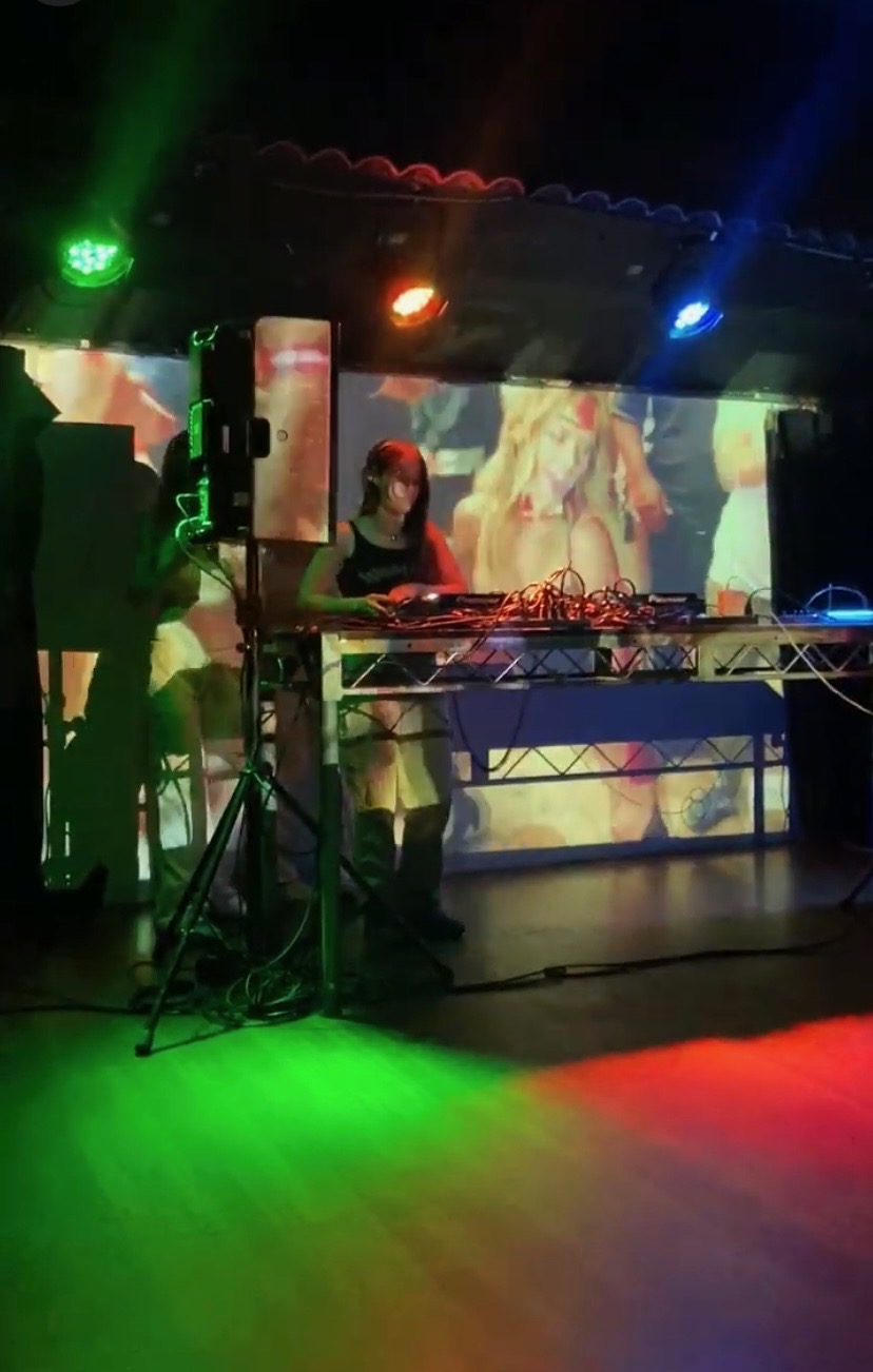 DJ at a booth with colorful lights and a large screen displaying abstract visuals in the background.
