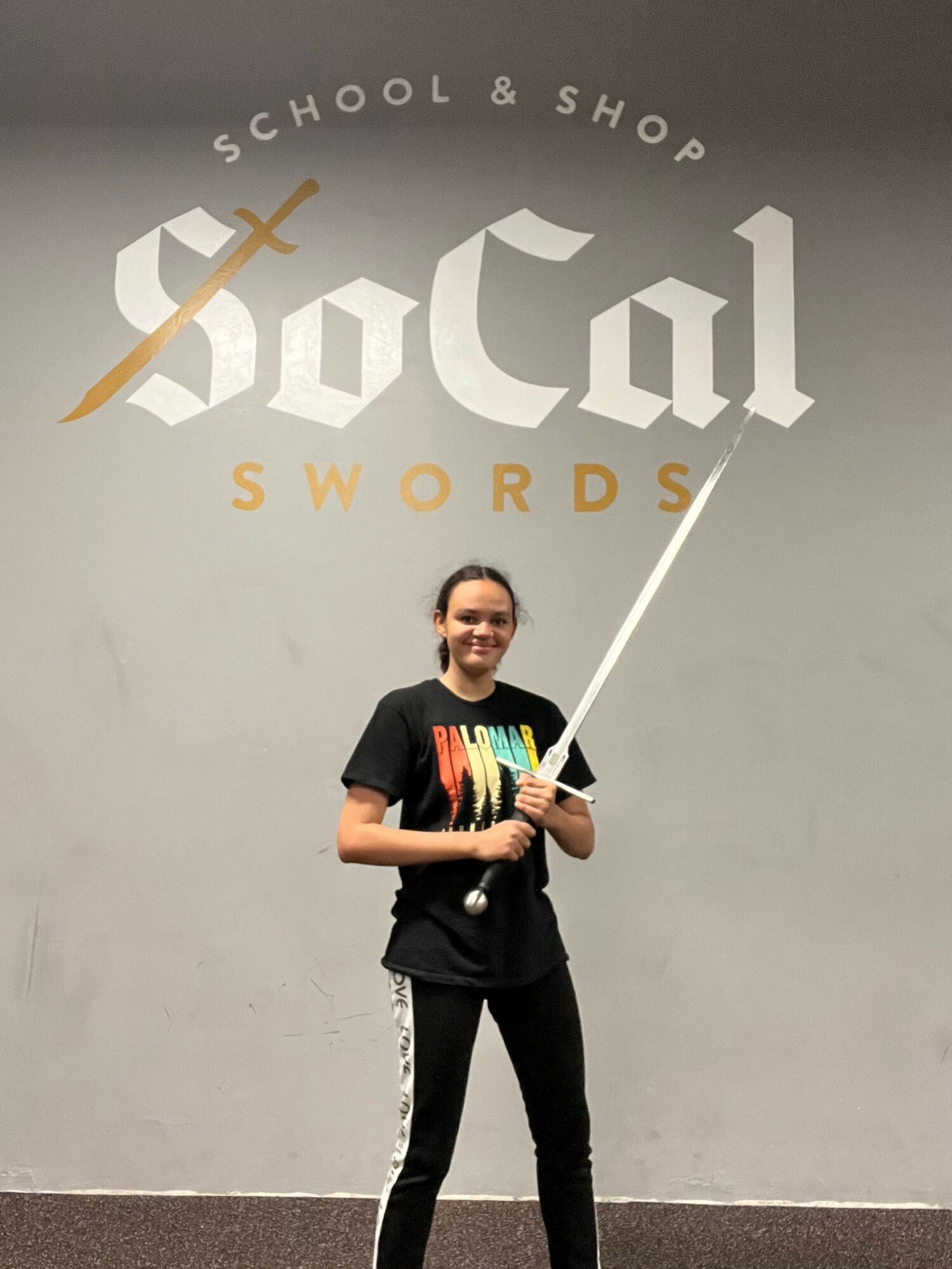 Person standing in front of a wall with a logo, holding a sword prop, smiling, wearing a black T-shirt and pants.
