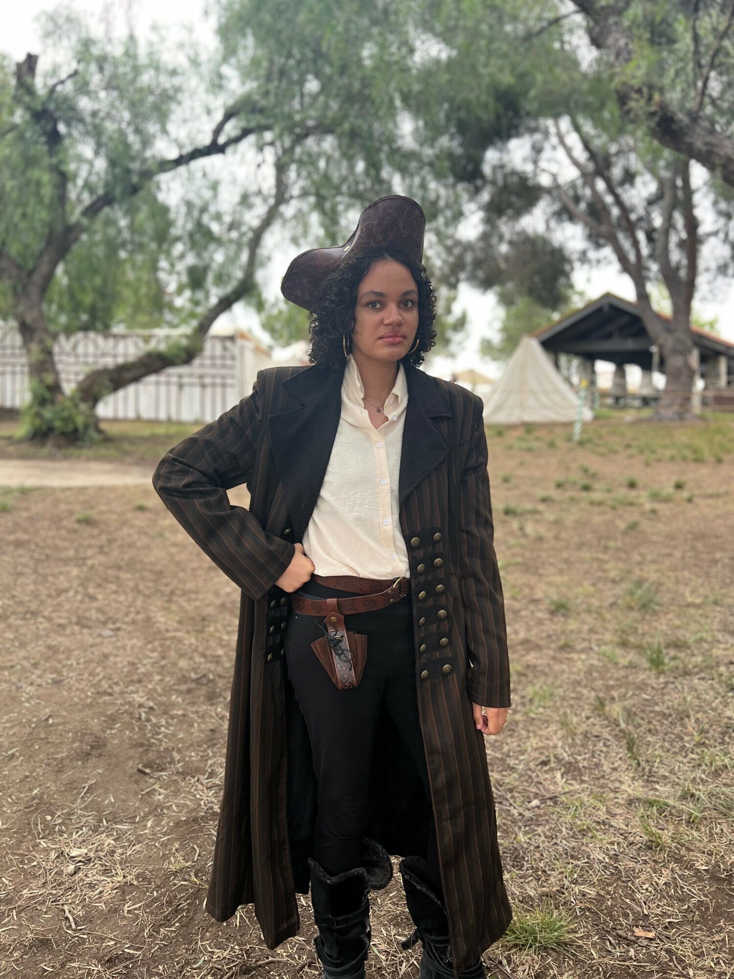 Person standing outdoors with trees and tents in background, wearing a long coat and dark pants.
