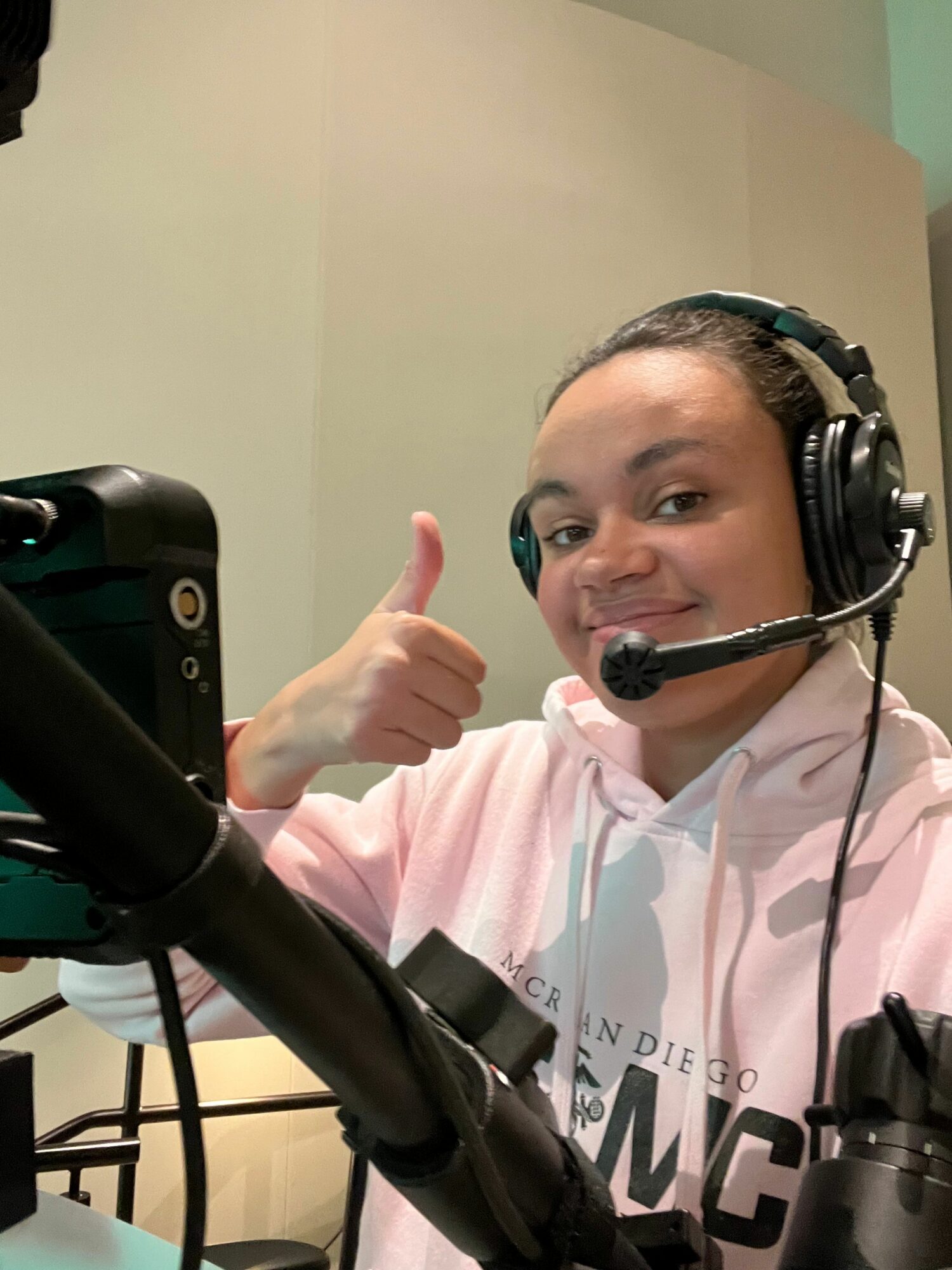 Young woman wearing headphones and a pink hoodie, giving a thumbs-up, smiling, in a recording studio.