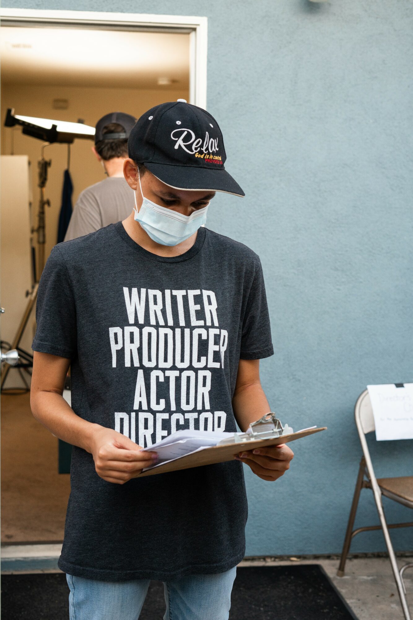 Person wearing a black cap, face mask, and a black T-shirt reading writer, producer, actor, director, holding a clipboard.