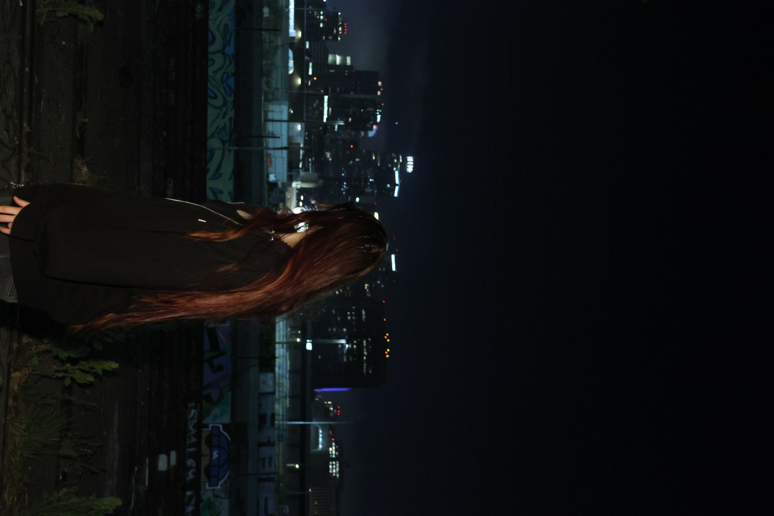 Person with long hair standing outdoors at night, city skyline with lights in background.