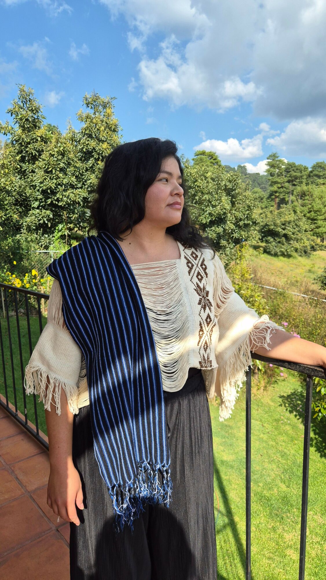 Woman standing outdoors on a balcony with trees and sky in background, wearing a cream top and striped scarf.
