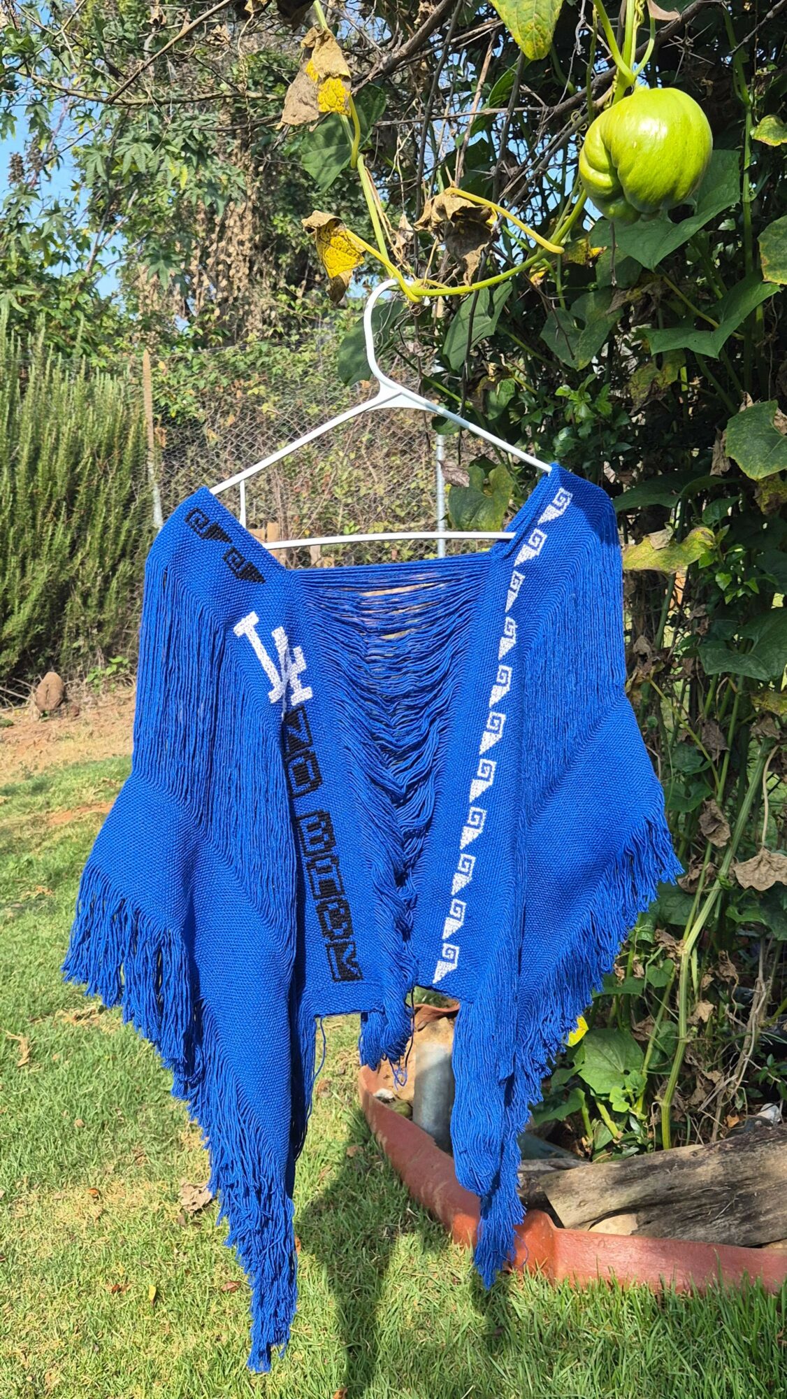 Blue knitted garment hanging on a white hanger outdoors with green foliage and a green fruit in background.
