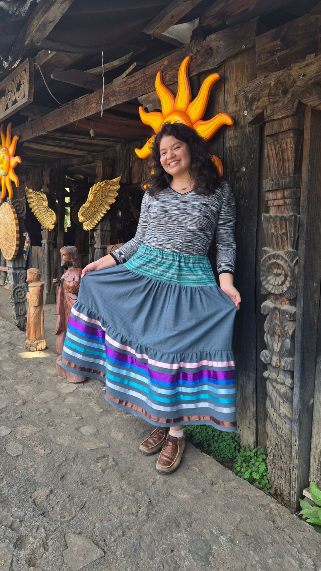 Woman in traditional dress with colorful skirt and headpiece, standing outdoors near wooden structures and decorative elements.
