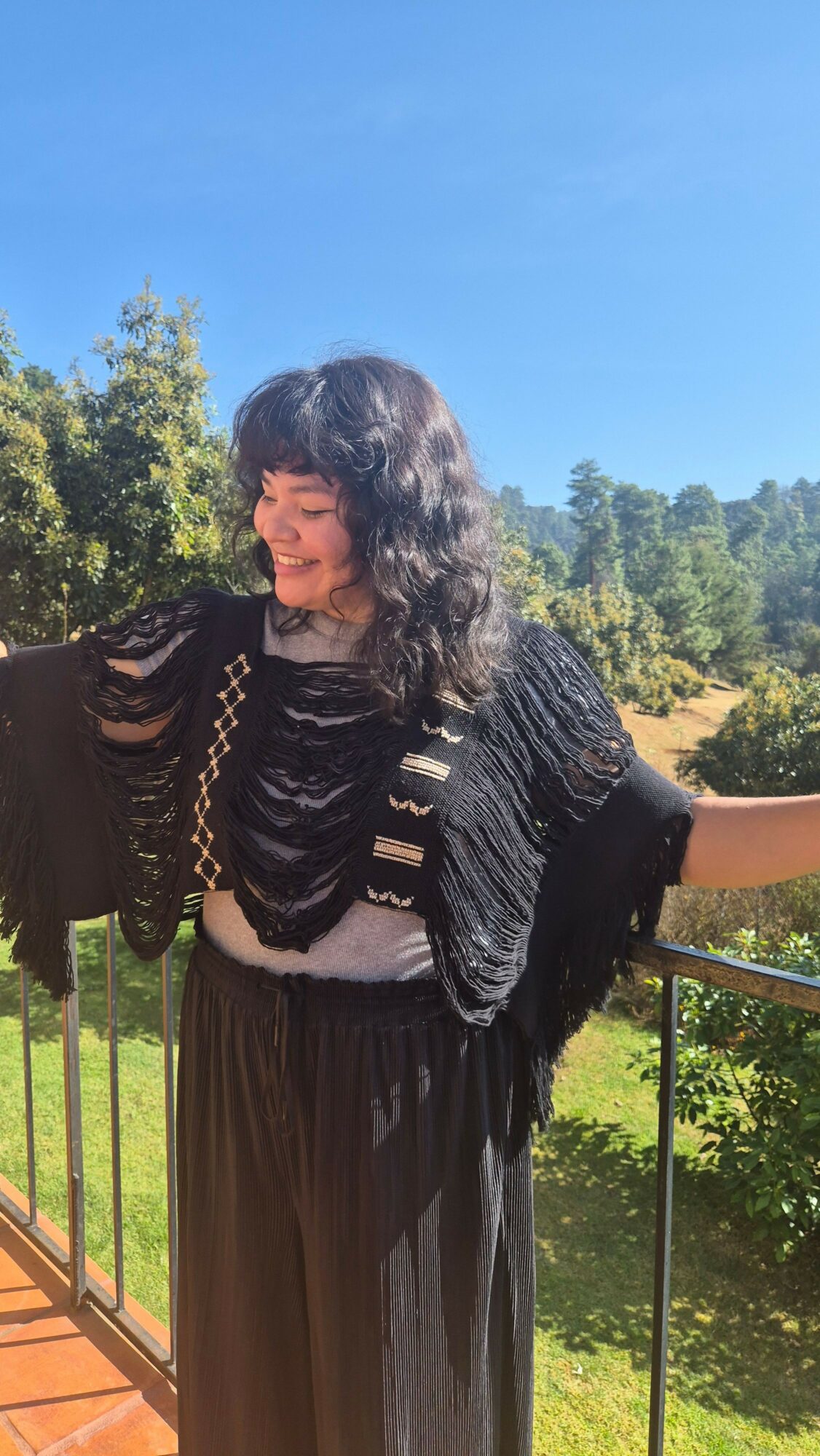 Young woman with curly hair smiling outdoors, wearing a black shawl and loose pants, with trees and blue sky in background.