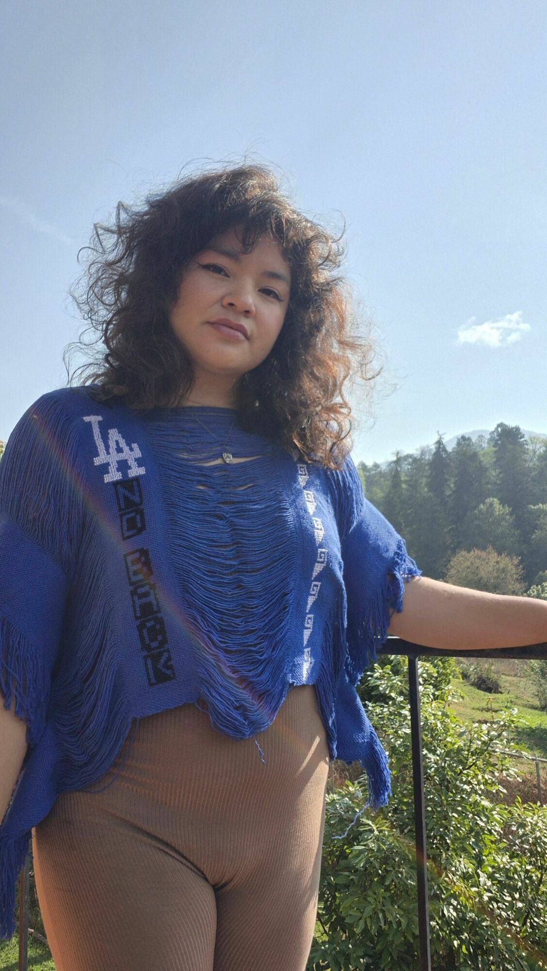 Woman with curly hair wearing a blue top and beige pants outdoors, with trees and sky in background.
