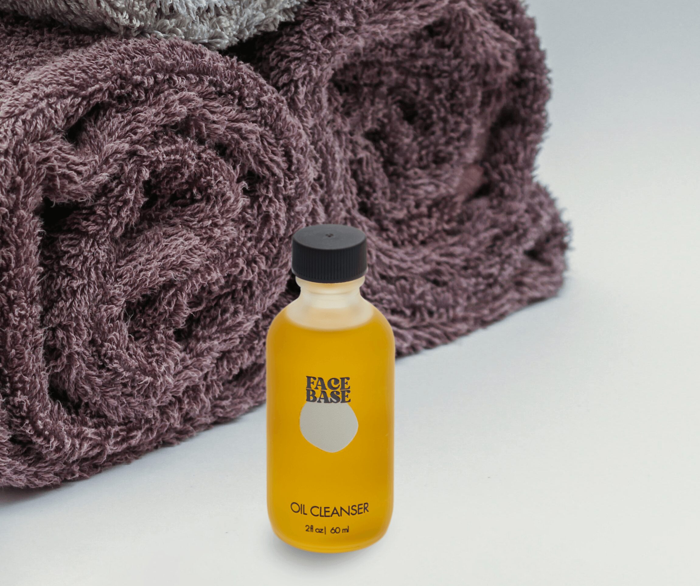 Small yellow bottle labeled 'Face Base Oil Cleanser' in front of folded purple towels.