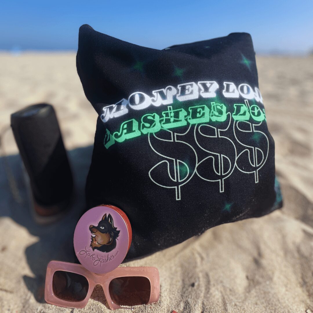 Black bag with green and white text, pink sunglasses, a pink circular object, and a black bottle on sandy beach.