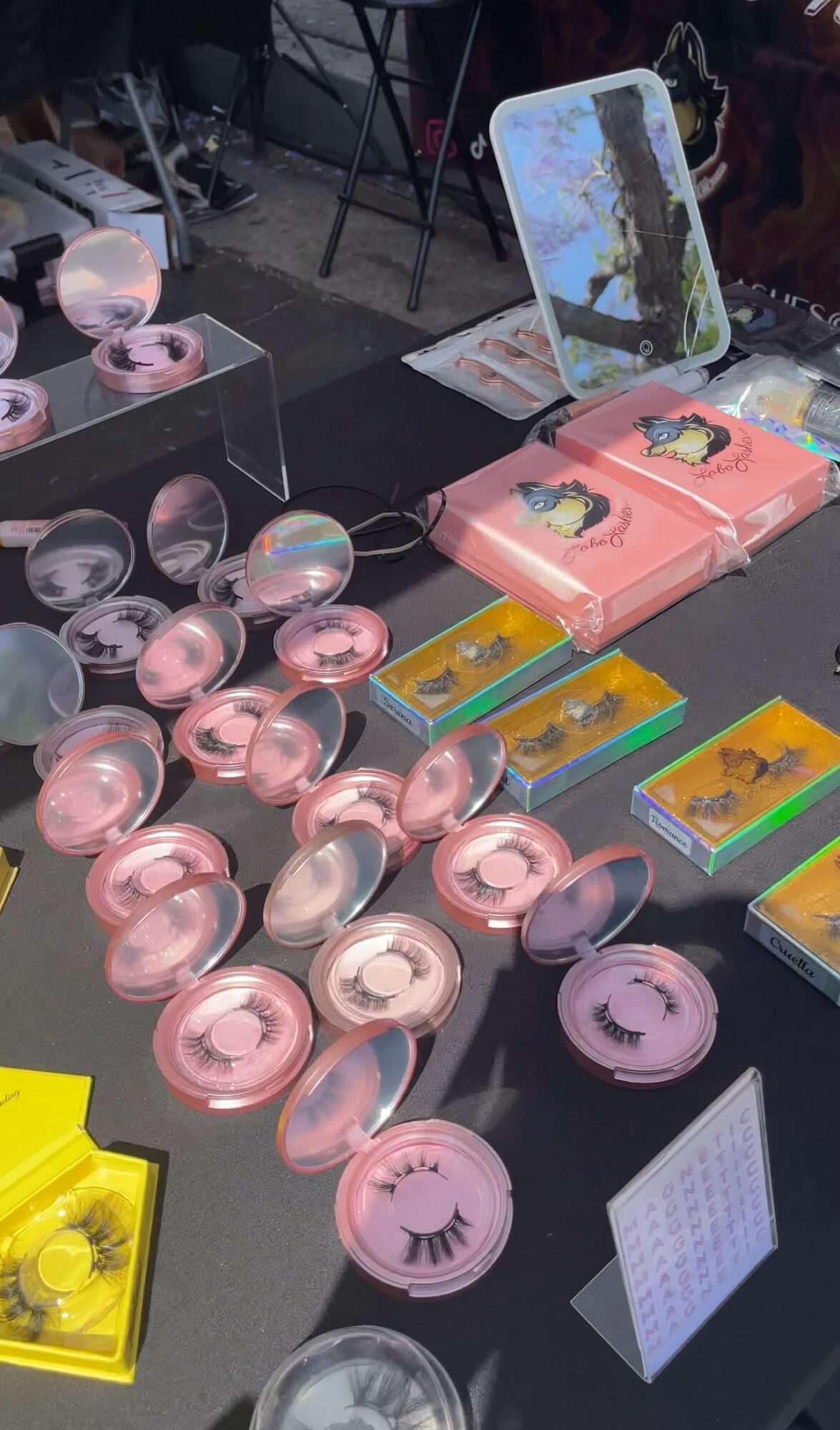 Table with pink and yellow packaged items, round pink containers, and a mirror with a stand, in a store setting.