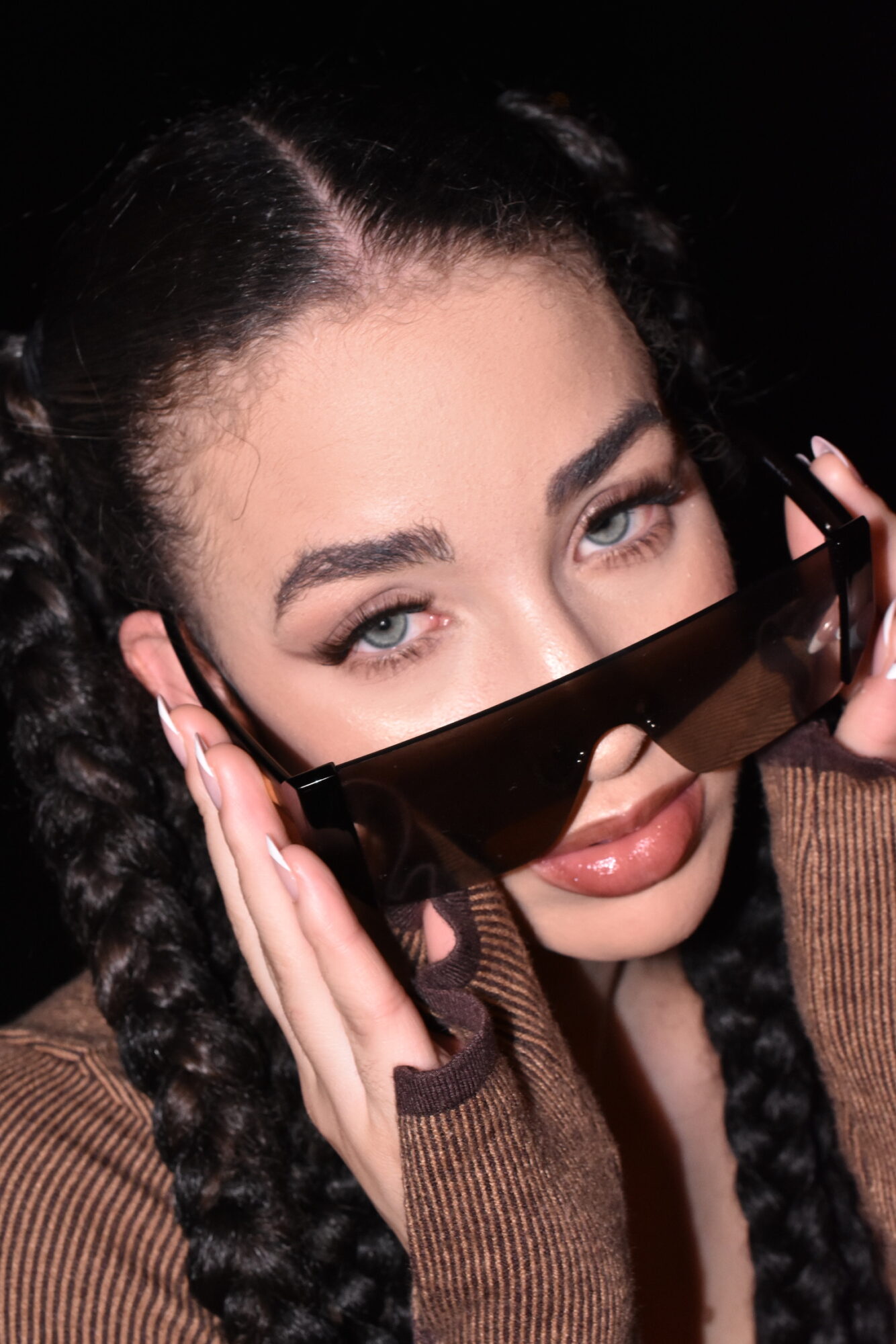 Woman with braided hair holding sunglasses near face, looking at camera, dark background.