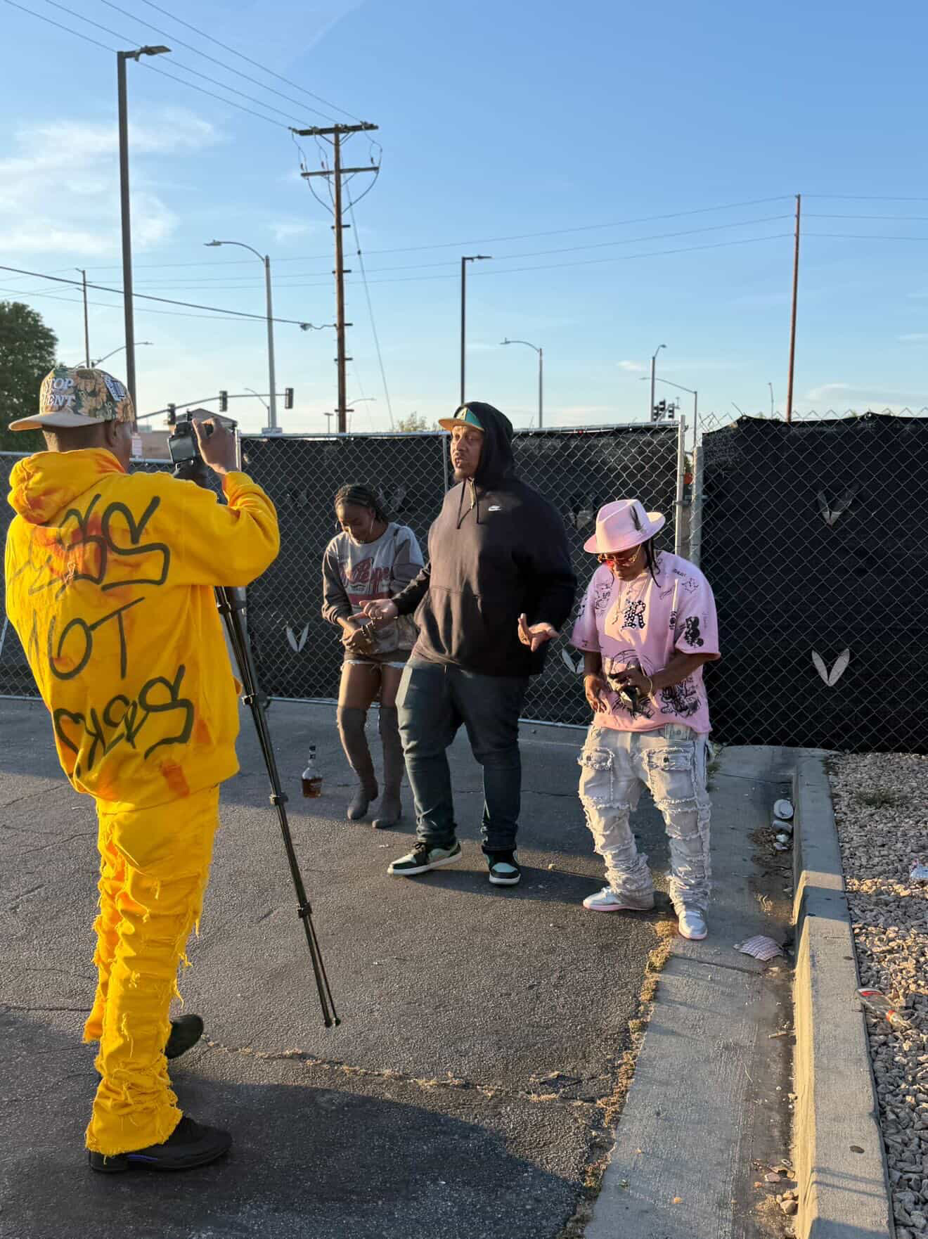 Four people standing on a street near a chain-link fence, with utility poles and streetlights in the background.