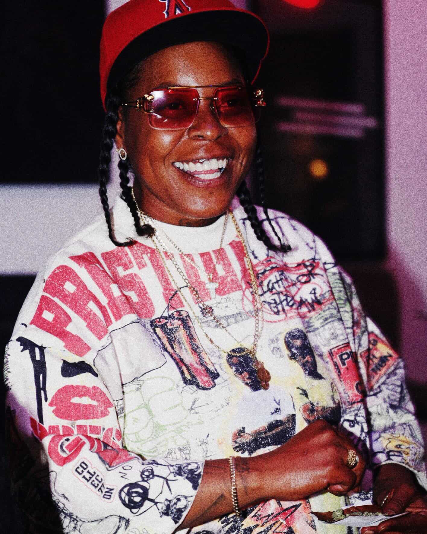 Smiling person wearing sunglasses, a red cap, and a colorful graphic jacket, holding a phone.