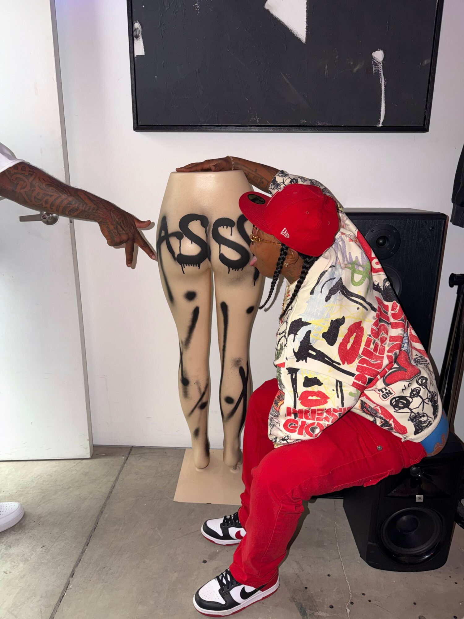 Person with red cap and colorful jacket crouches near a mannequin with graffiti-style markings, in a room with a speaker and wall art.