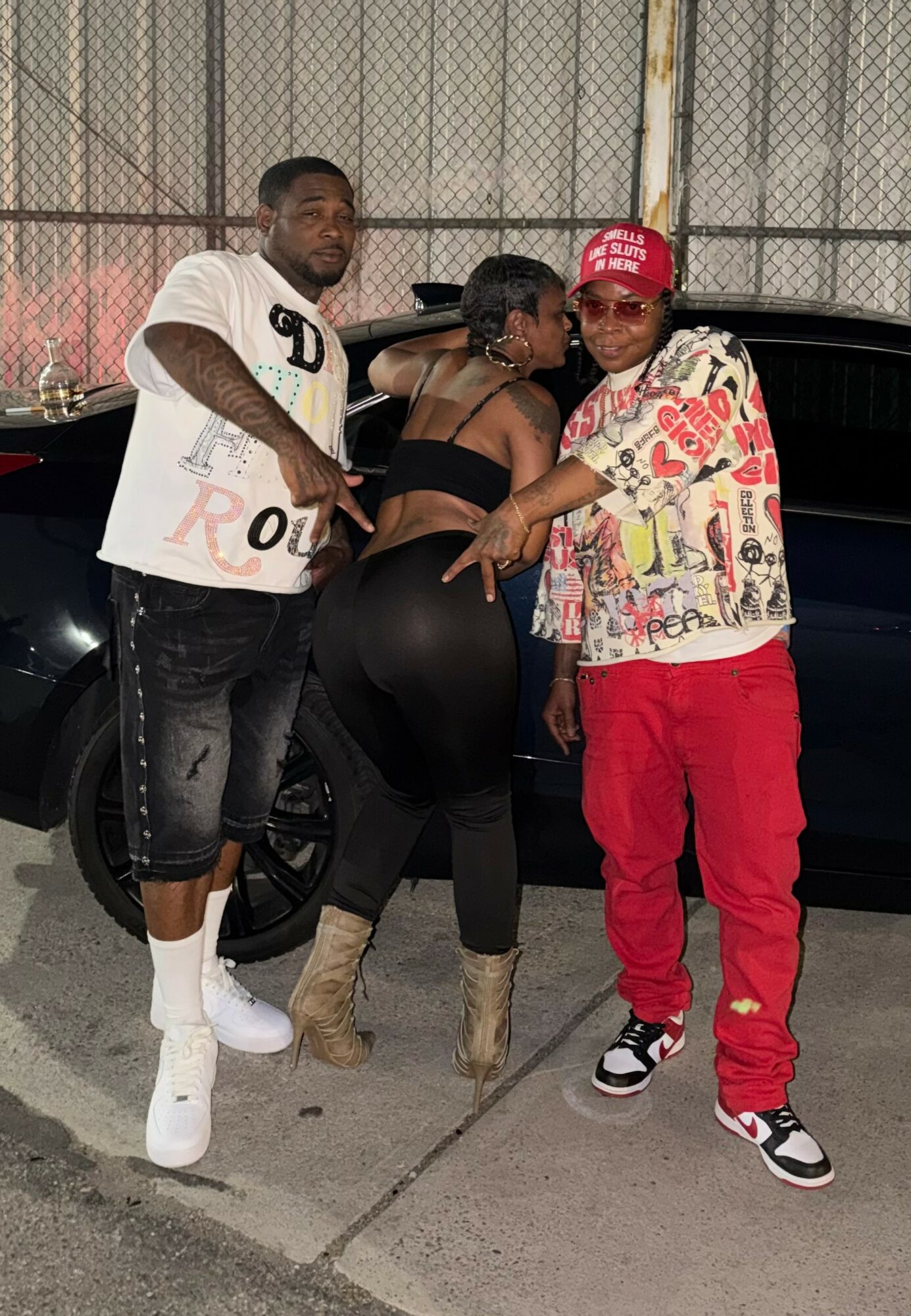 Three people standing outdoors near a black car, with a chain-link fence in the background. One person wears a white shirt, another a black top, and the third a colorful shirt and red pants.