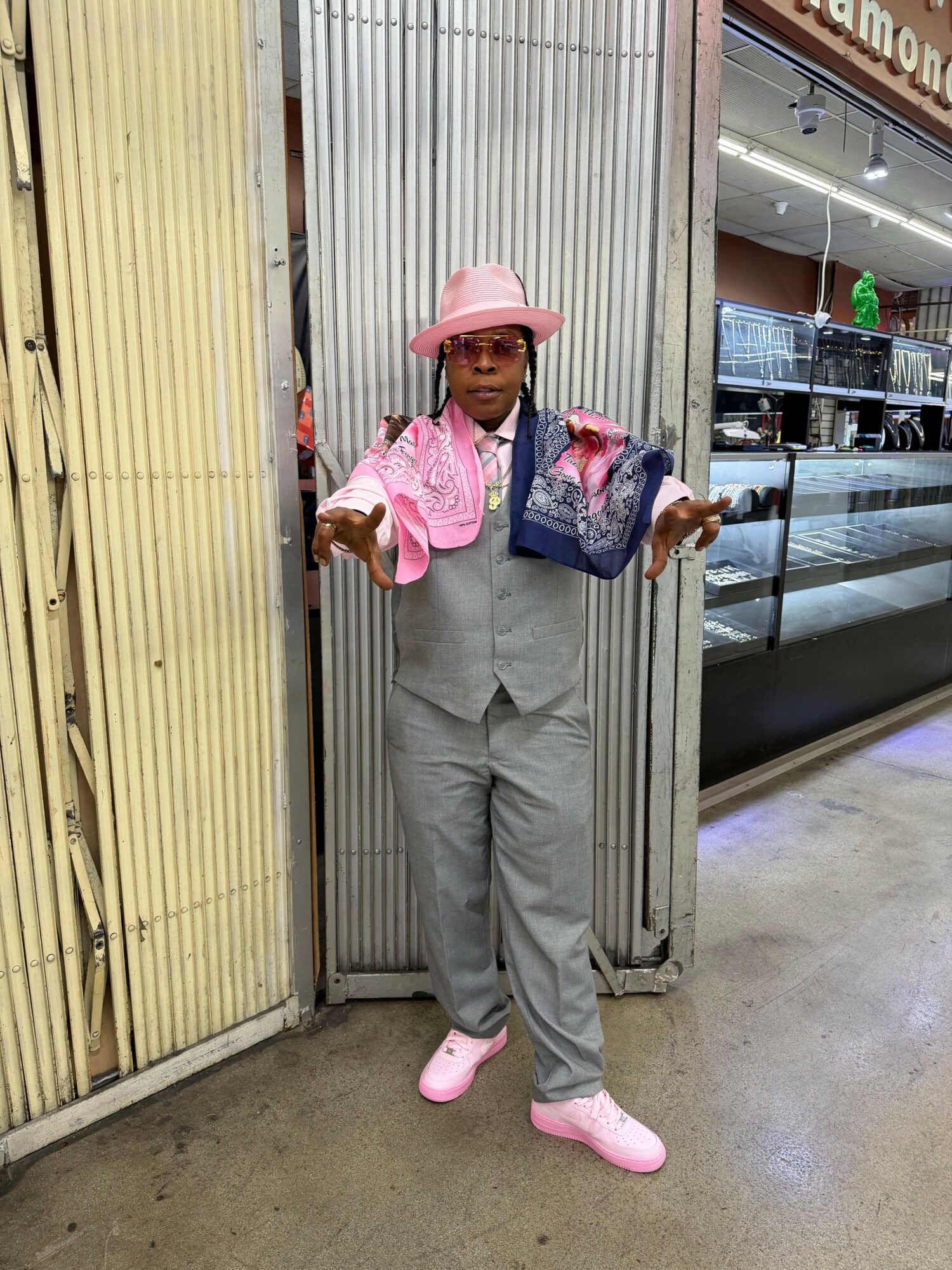 Person in gray suit with pink accessories and hat, standing in front of a metal wall inside a store.