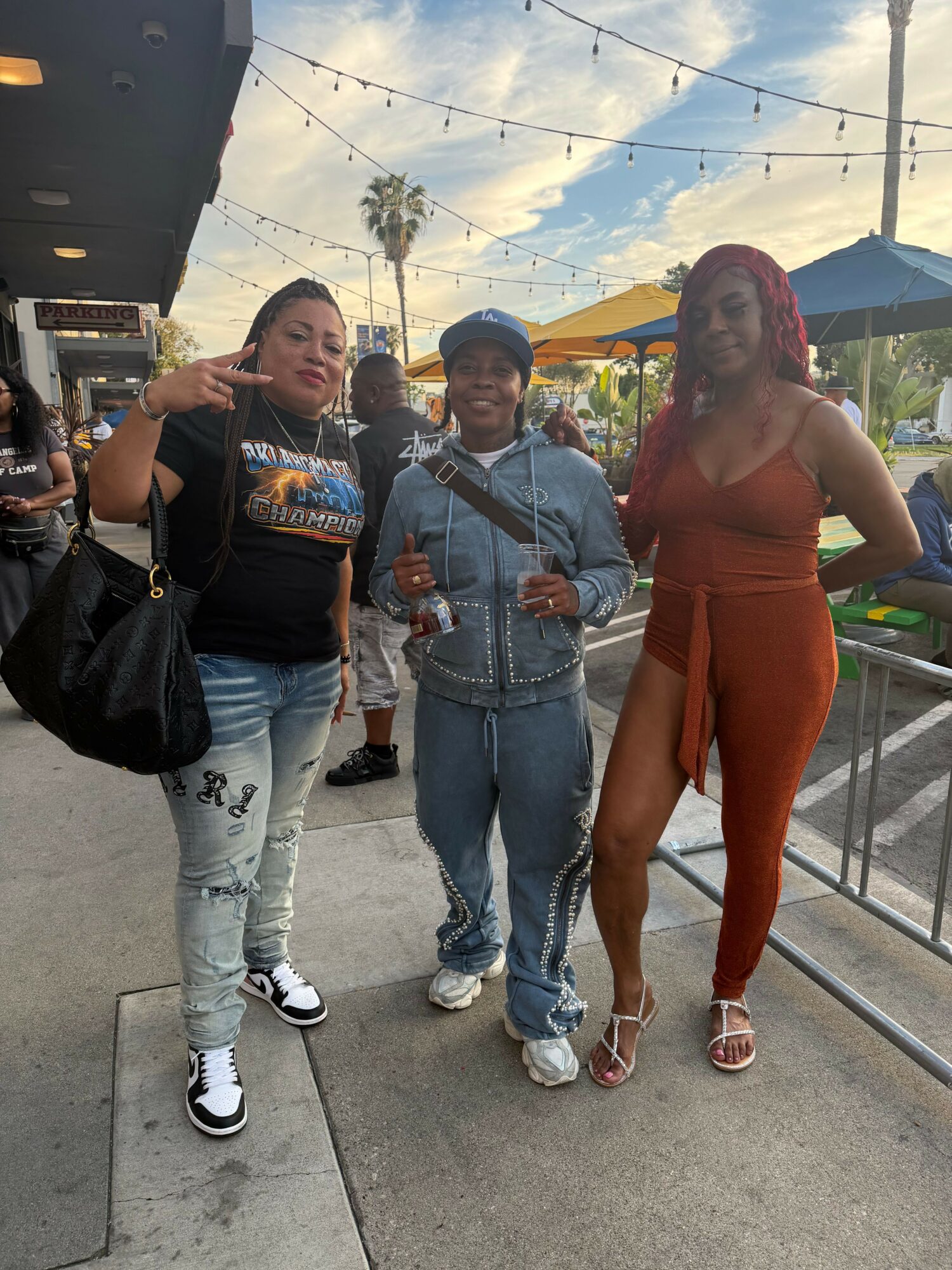Three women standing outdoors, smiling, with colorful umbrellas and string lights overhead, in a lively outdoor setting.