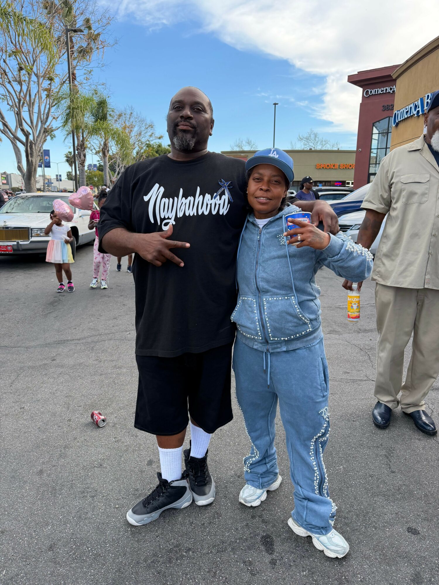 Two people standing outdoors, one making a peace sign, with a group and trees in the background.