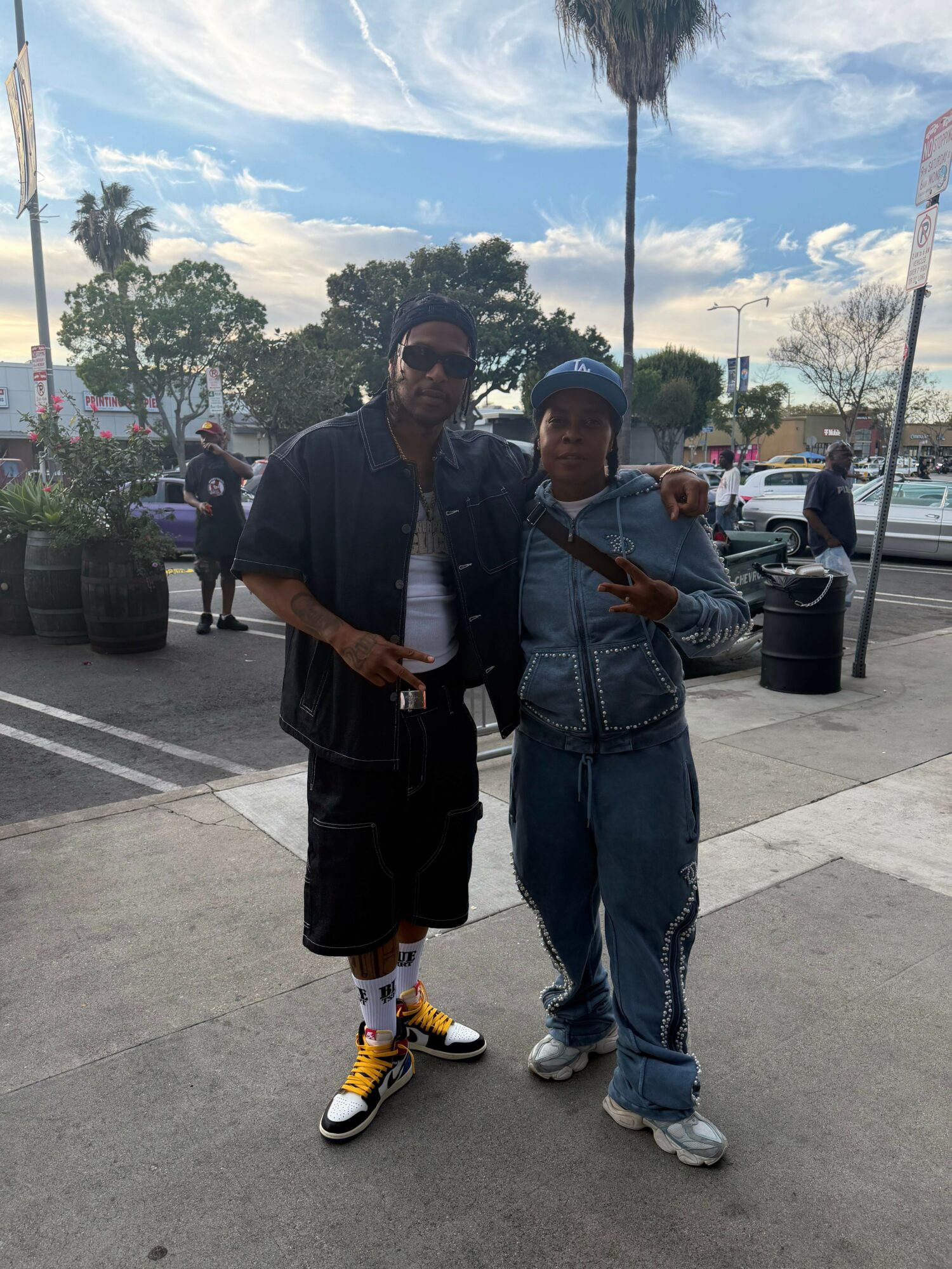 Two men standing outdoors in a parking lot with palm trees and cars in background.