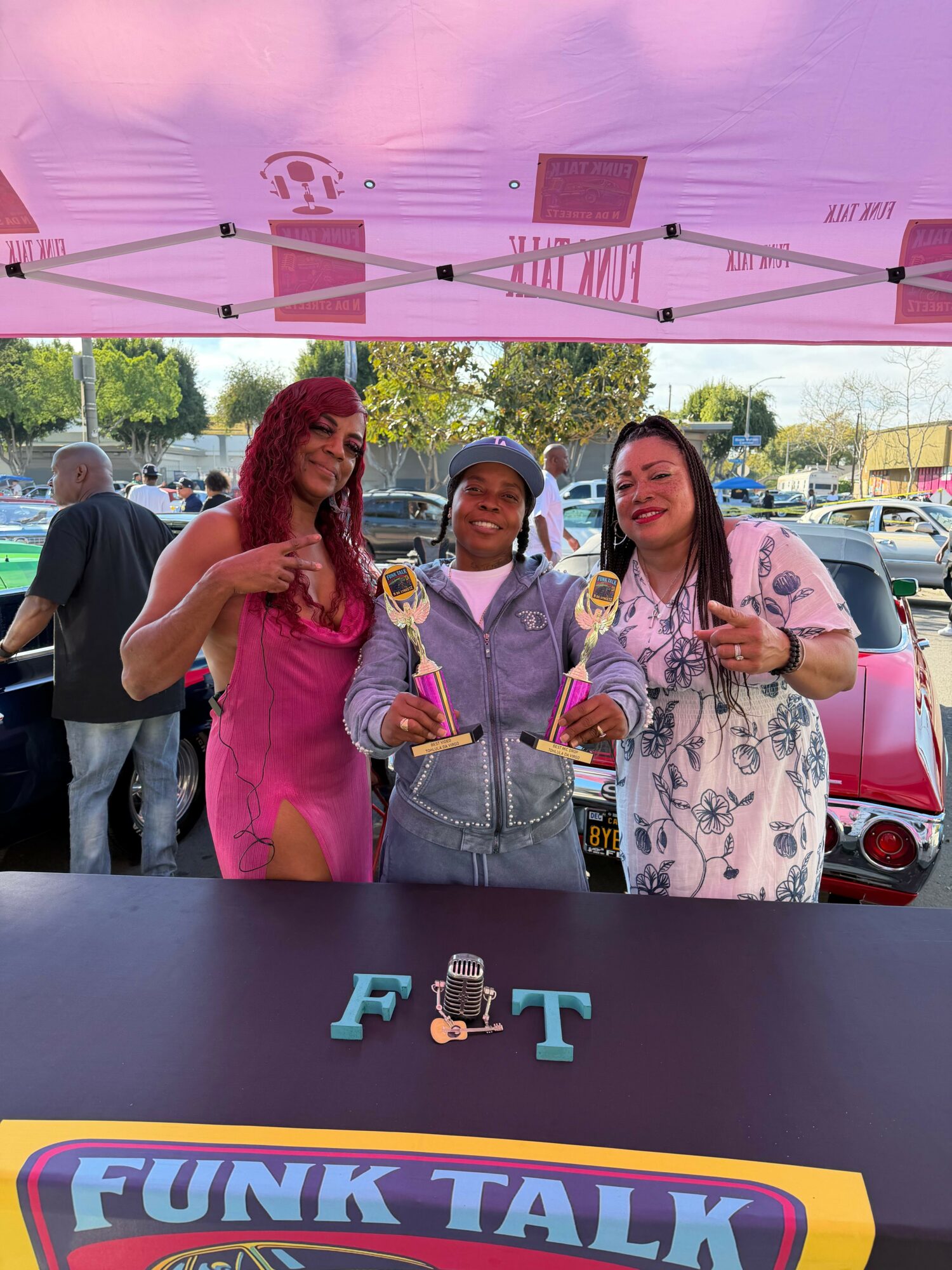 Three women stand behind a table with colorful letters and a small figure, smiling outdoors under a pink canopy.
