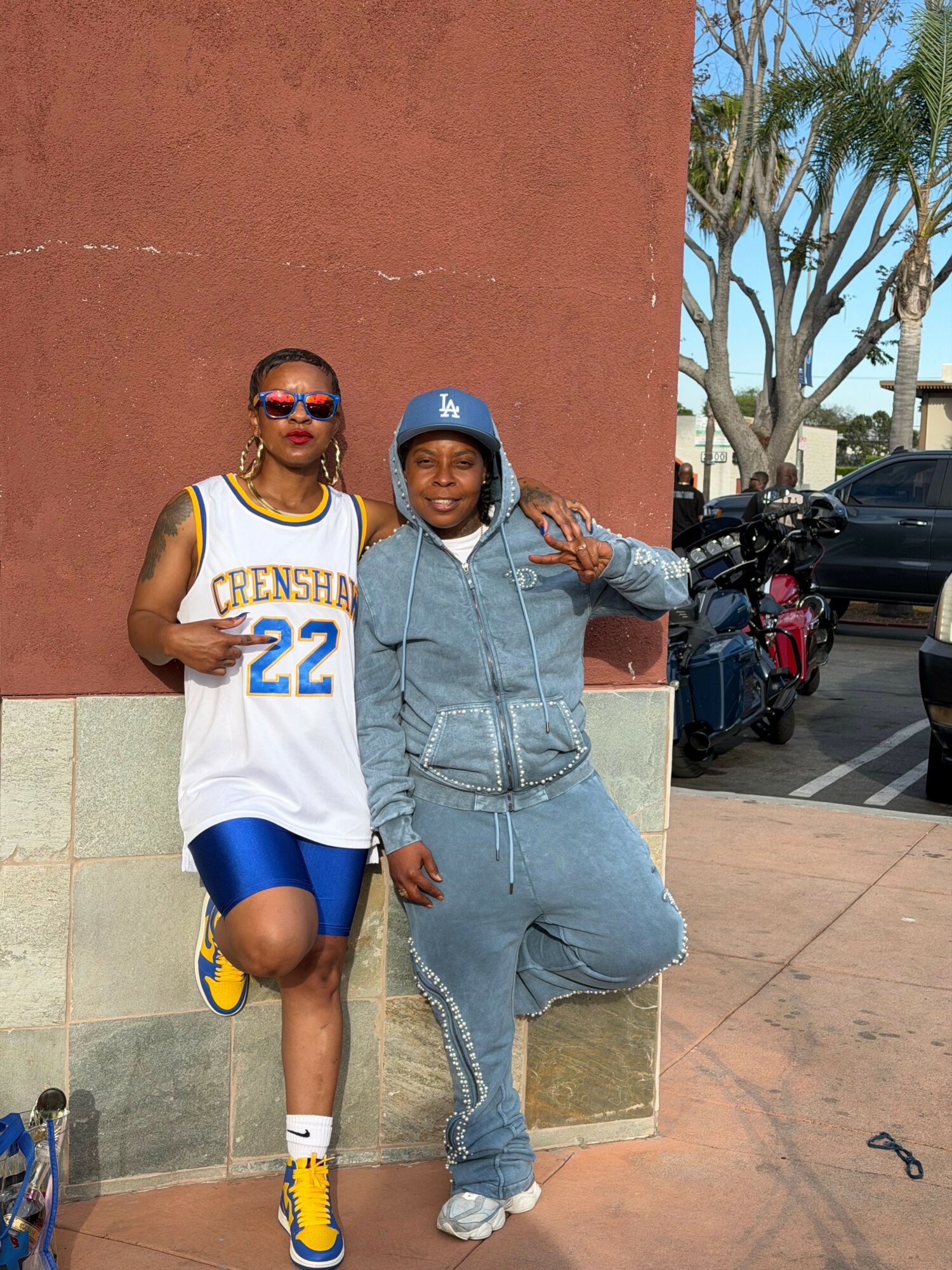 Two women standing against a red wall, one in a basketball jersey and the other in a hoodie, making a peace sign.