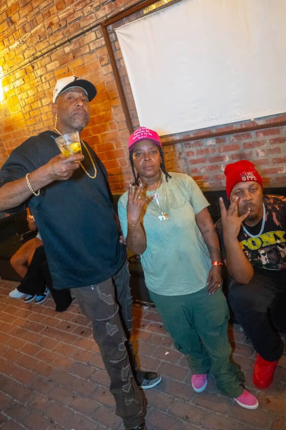 Three people standing against a brick wall, one holding a drink, wearing casual clothing and accessories.