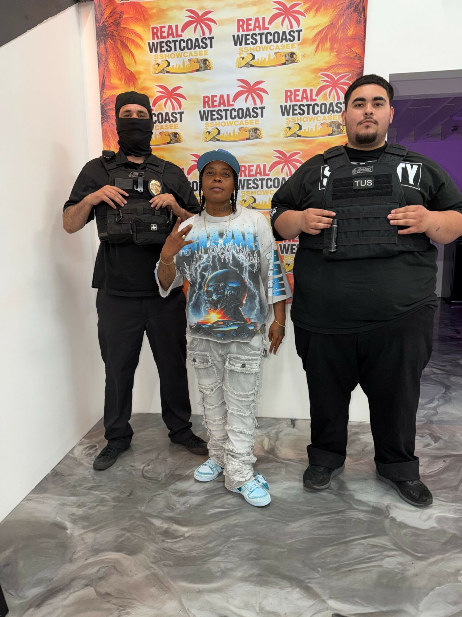 Child standing between two police officers in front of a Real Westcoast backdrop, making a peace sign with one hand.