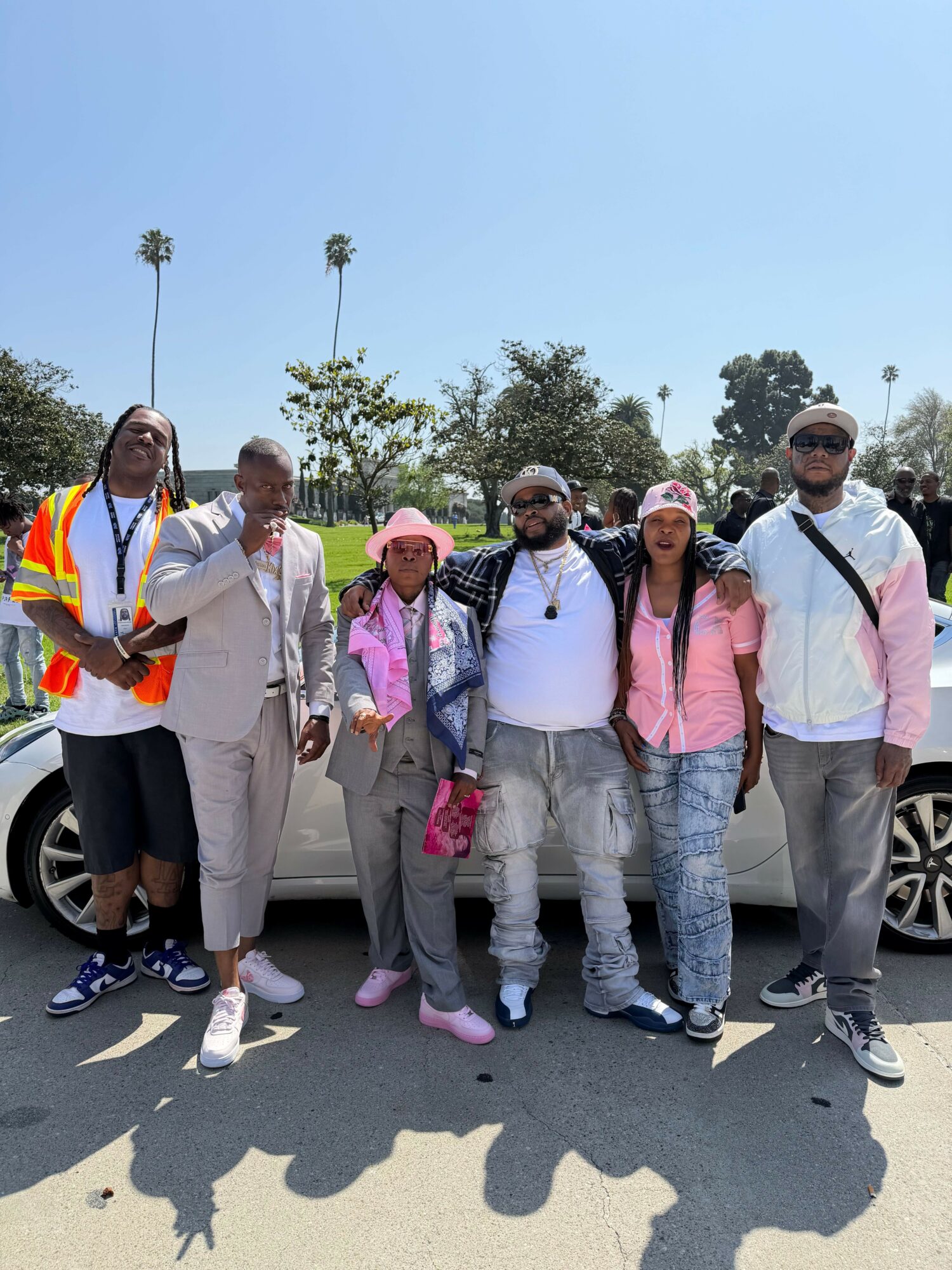 Group of six people standing outdoors in a parking lot, with trees and blue sky in background.