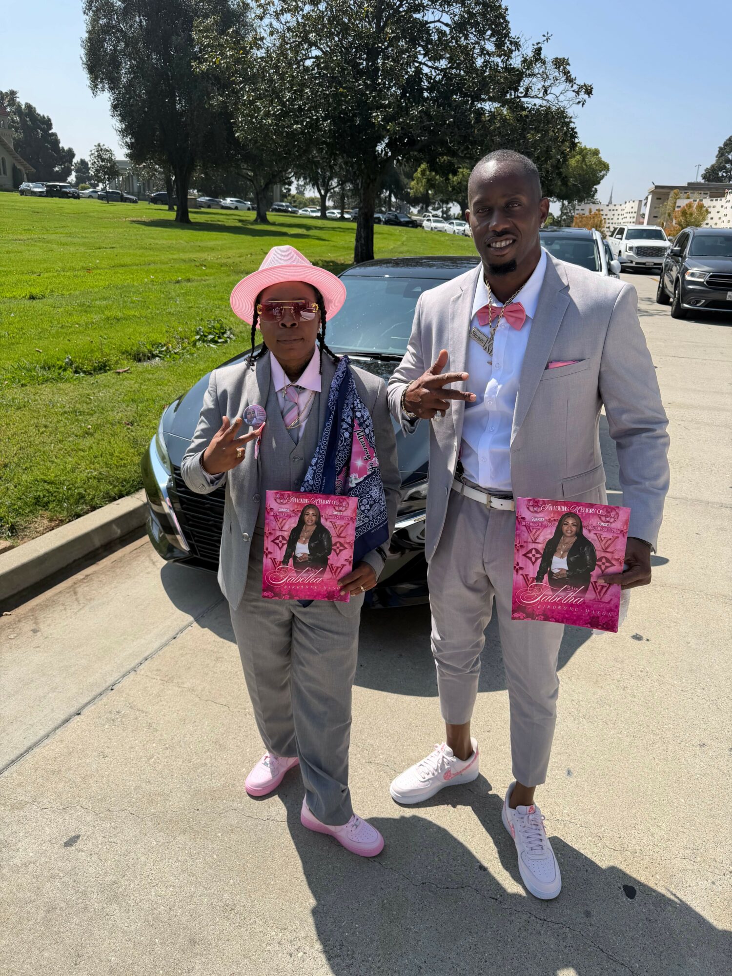 Two people standing outdoors on sidewalk holding pink bags with a woman wearing a pink hat and sunglasses, man in a light suit.