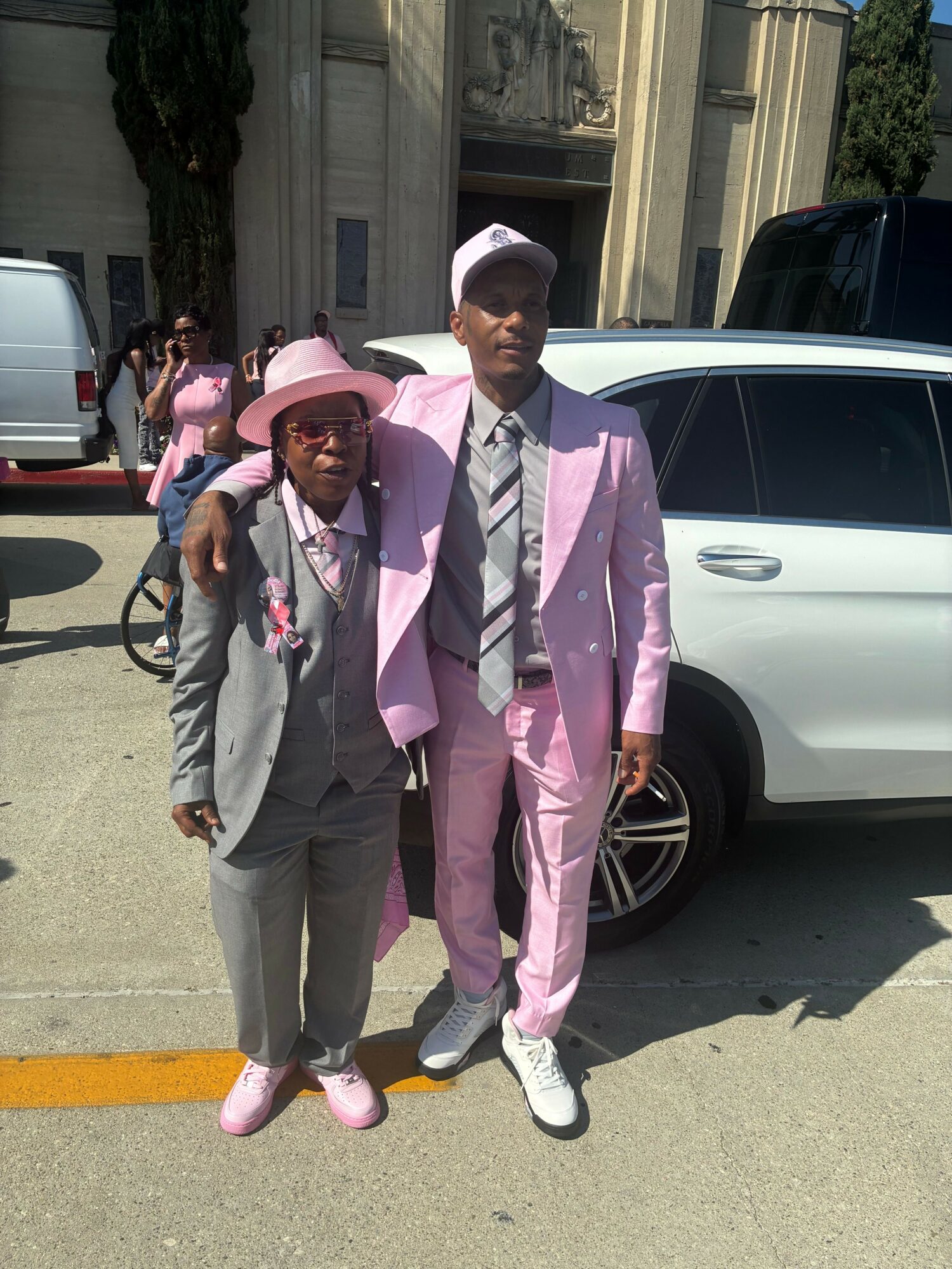 Two people standing outdoors in front of a white car, dressed in pink suits and hats, with a building in background.
