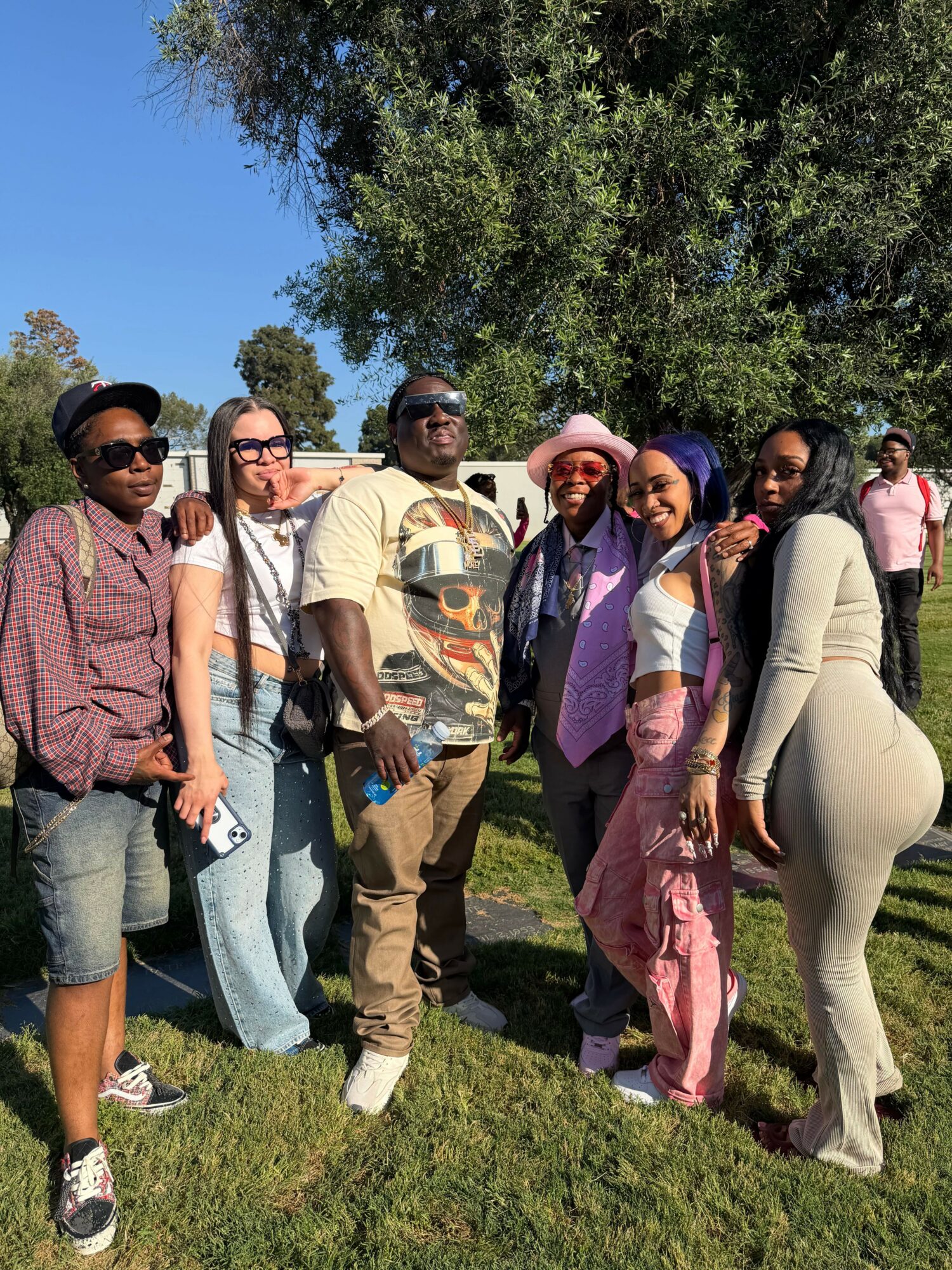 Group of six people standing outdoors on grass with trees and blue sky in background.