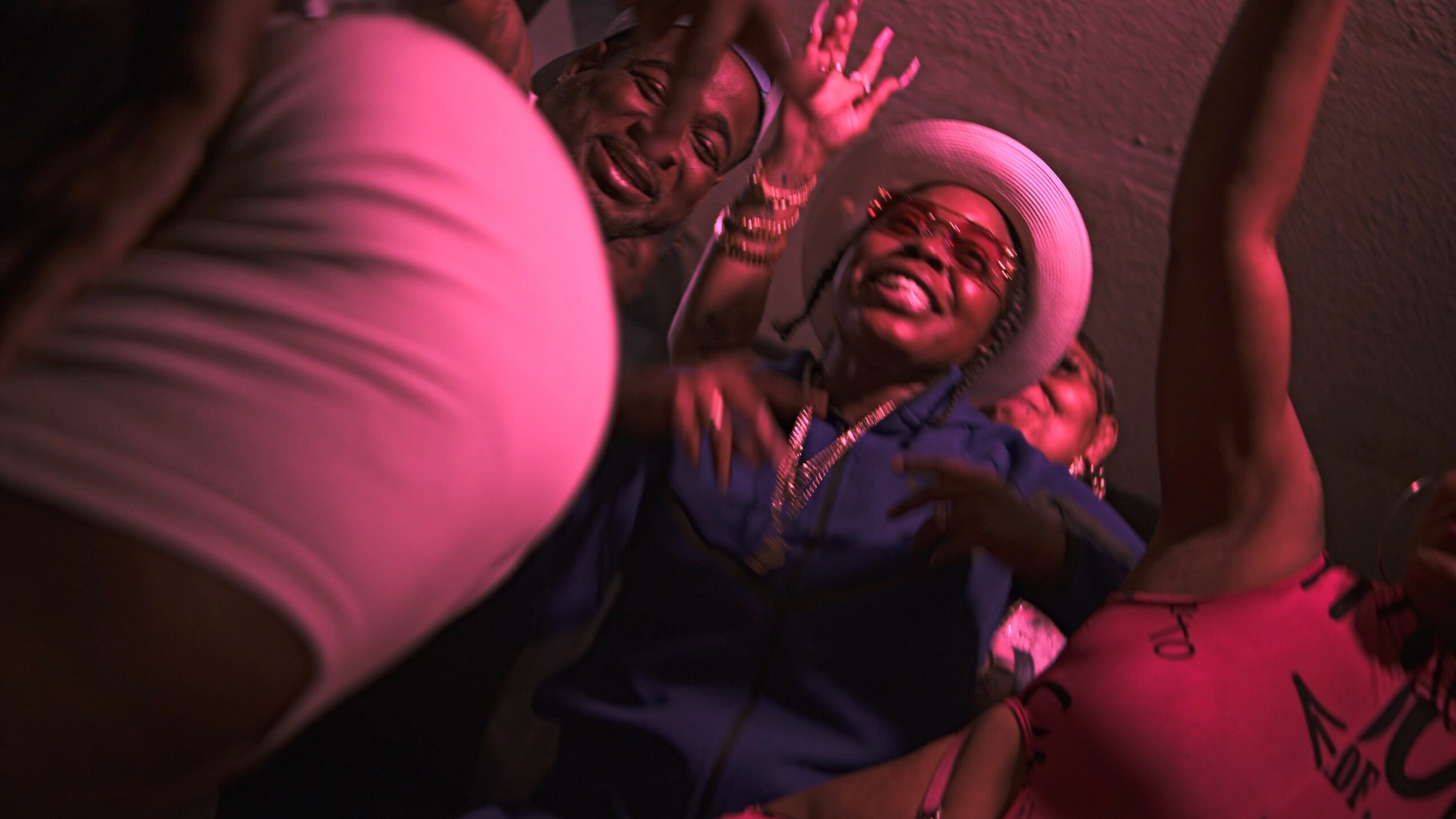 Group of people smiling and dancing indoors, with one person wearing a pink hat and others raising hands.