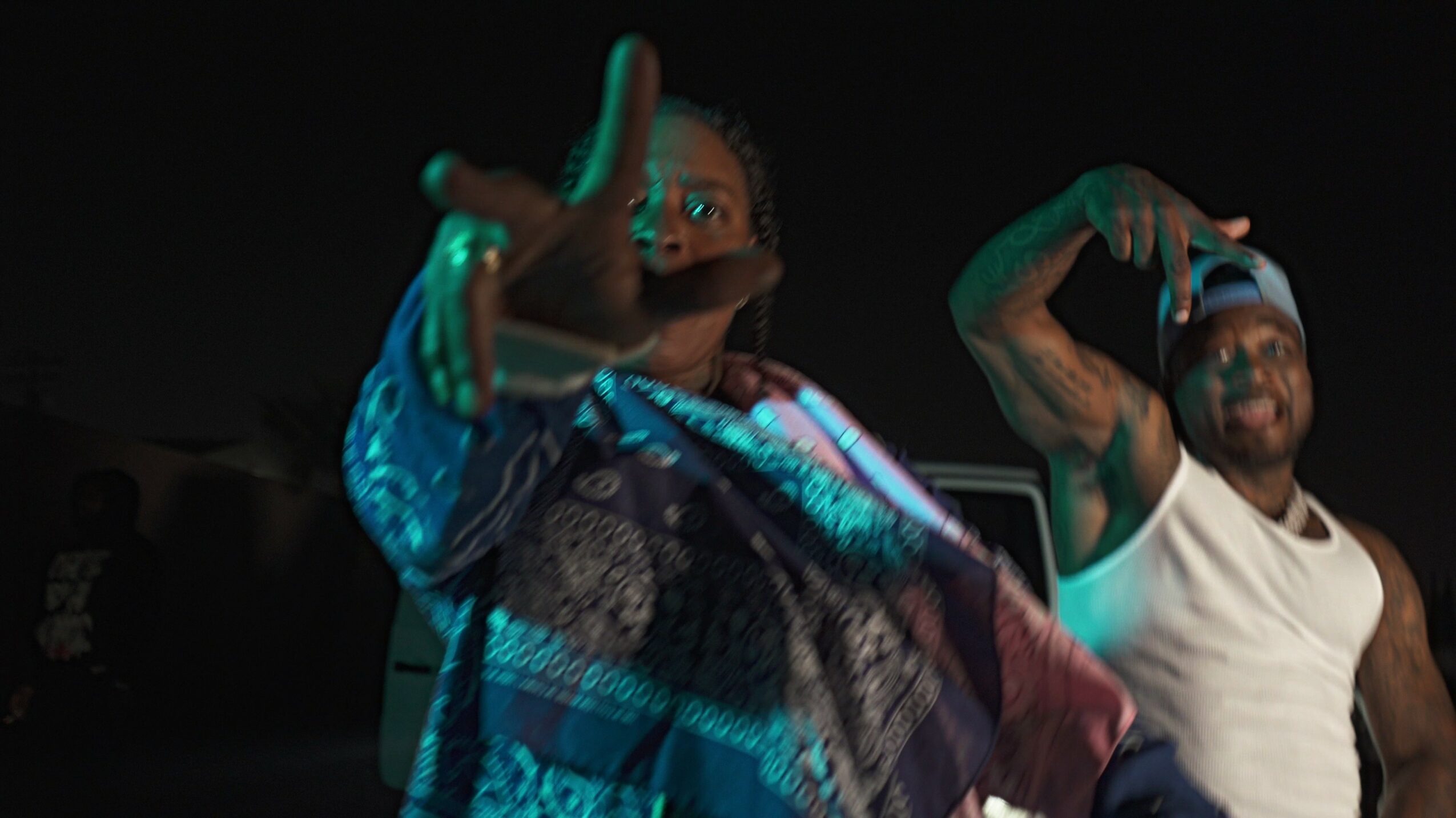 Two men in casual clothing pose with hand gestures in a dark setting.