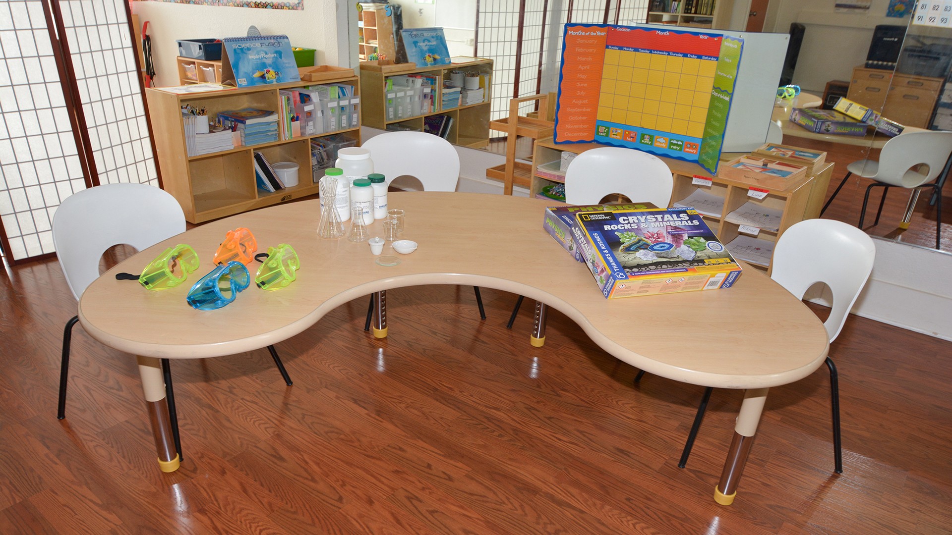 Curved wooden table with chairs, books, water bottles, and toys in a room with bookshelves and a colorful chart.