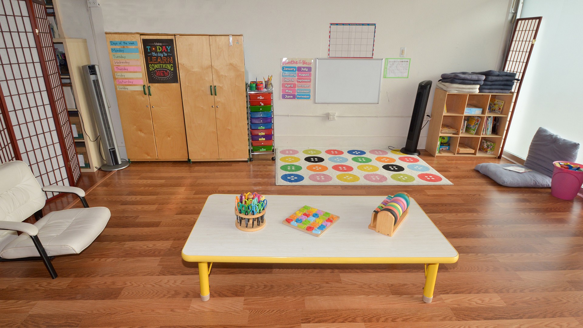 Classroom with a white table, colorful mats, storage units, and chairs on a wooden floor.