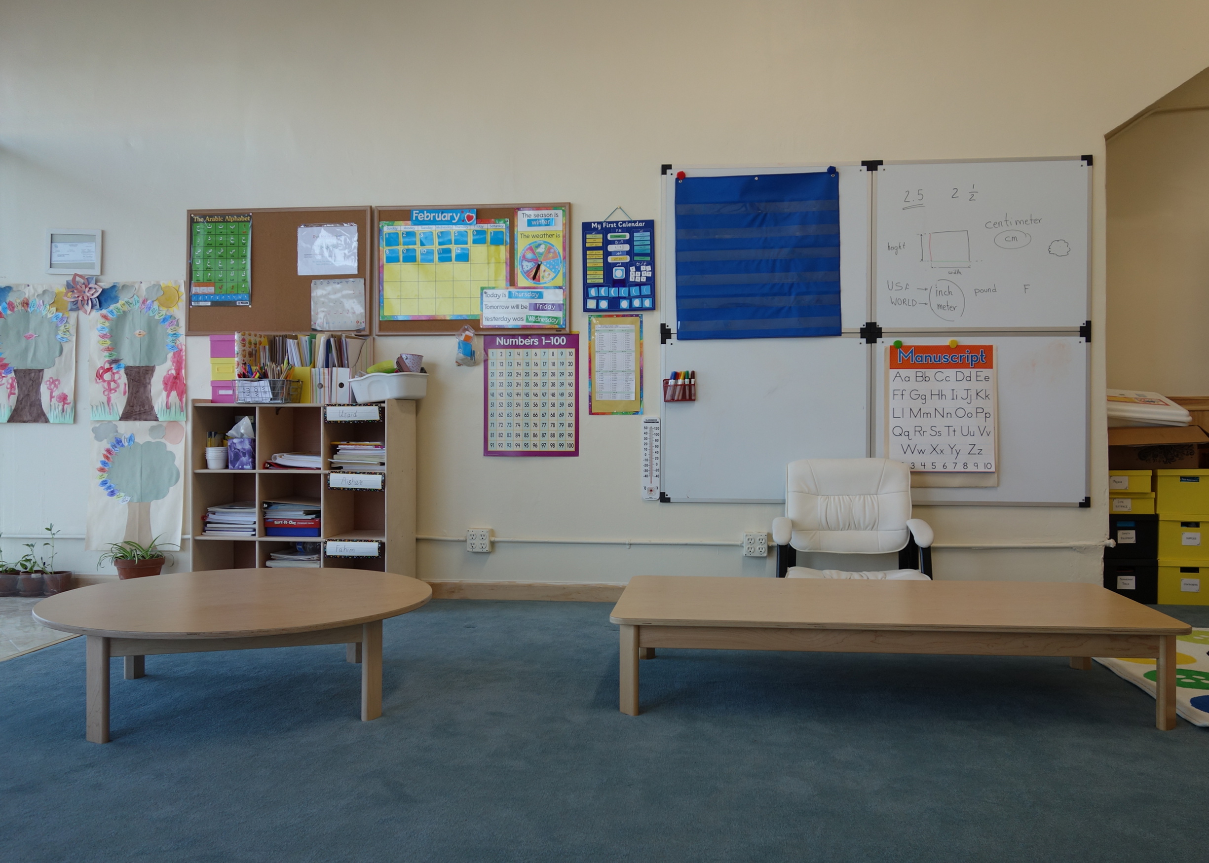 Classroom with whiteboard, bulletin boards, shelves, a chair, and a bench on a blue carpet.