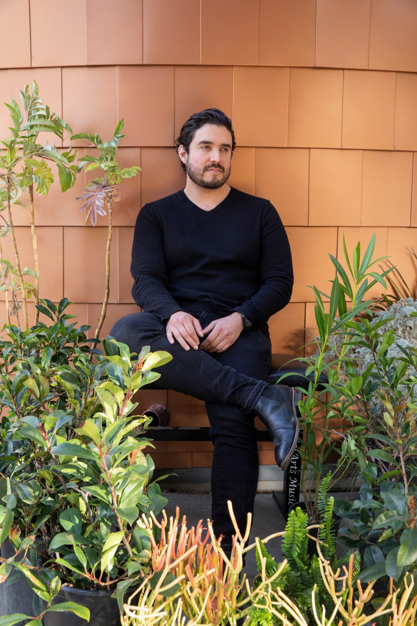 Man with beard and dark clothing sitting on a bench surrounded by green plants, against a wooden wall background.
